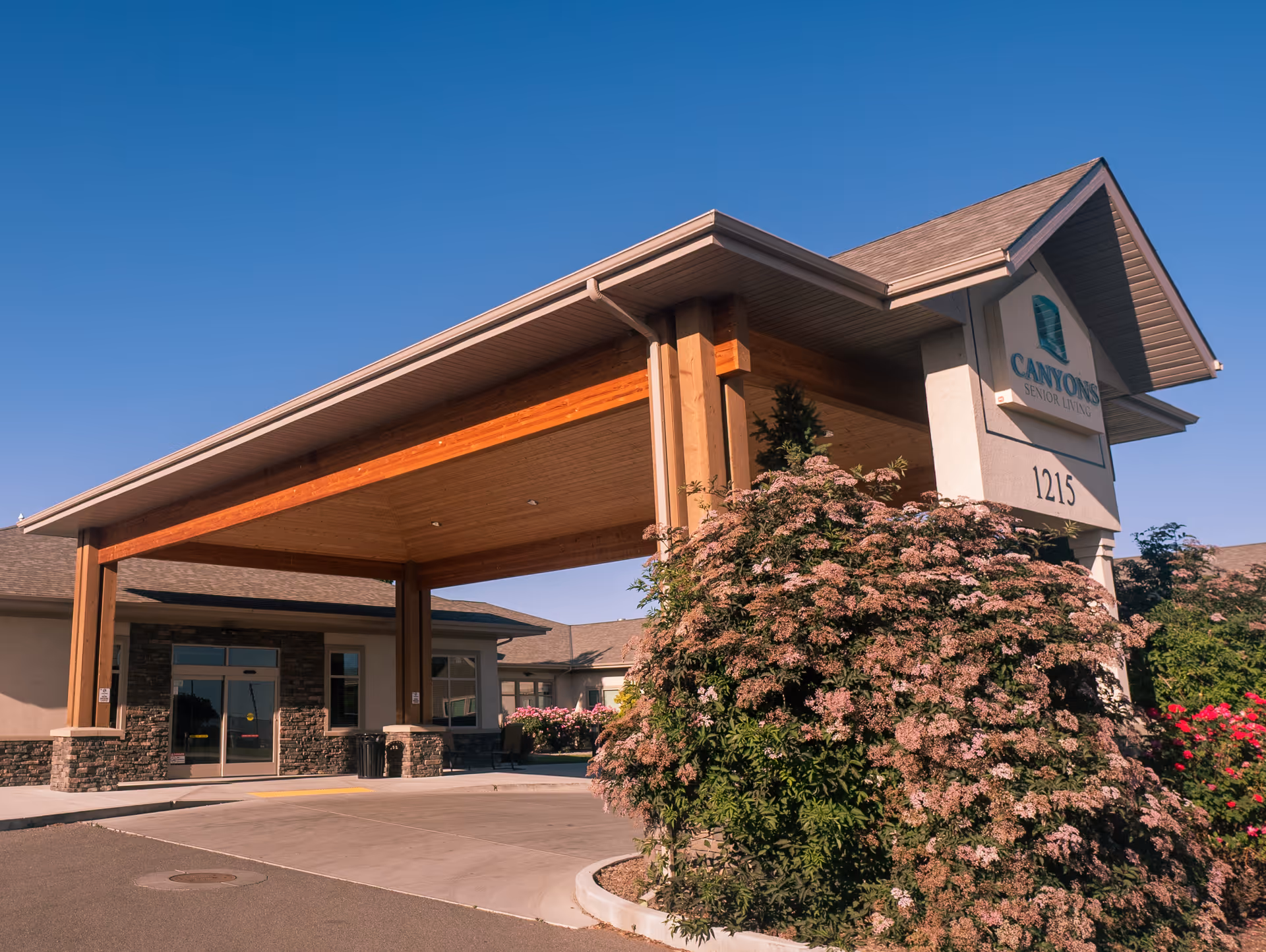 Exterior view of the entrance to Canyons Senior Living facility with a covered drop-off area supported by wooden beams, surrounded by bushes and flowers under a clear blue sky.