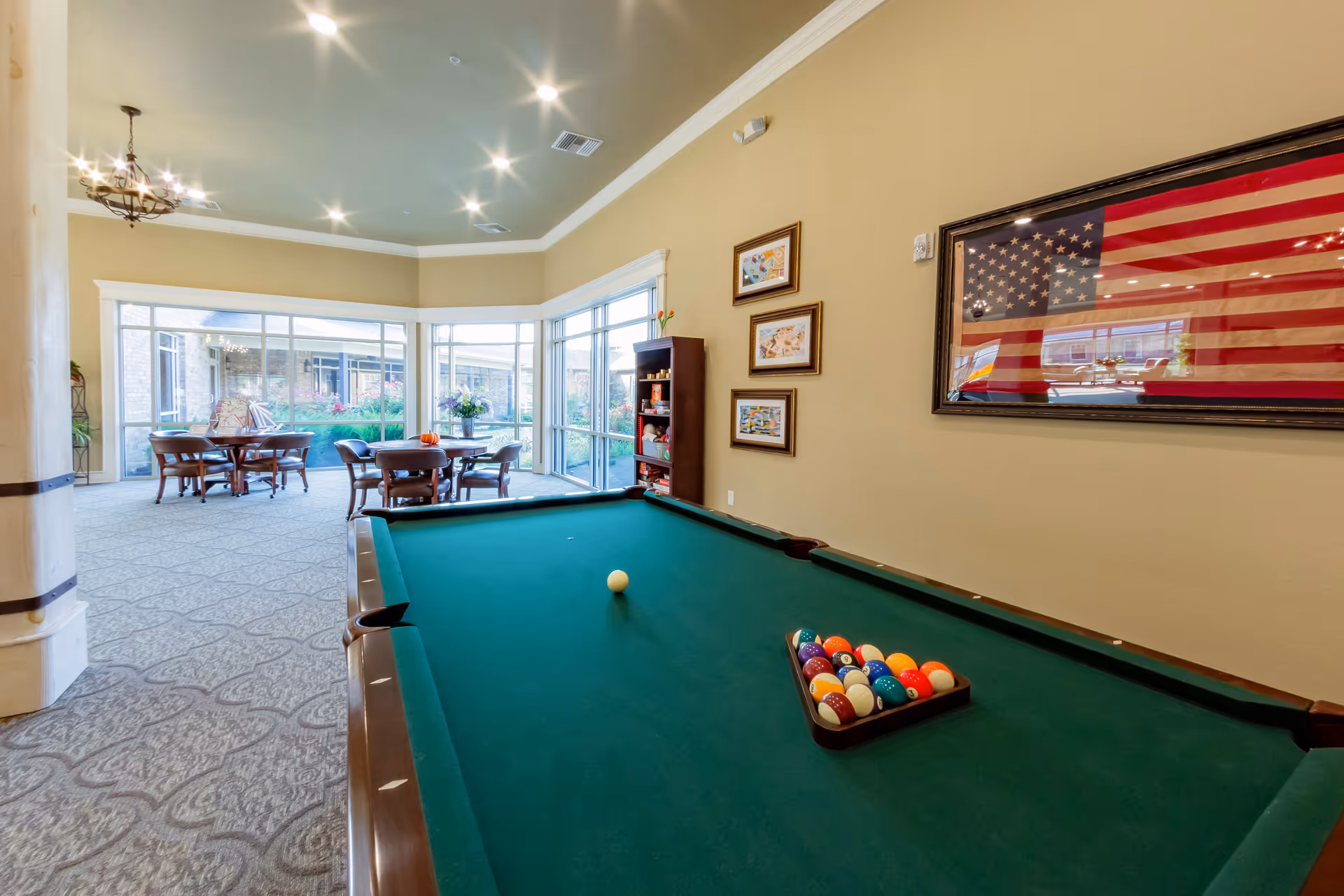 Interior view of a recreational room in a senior living facility featuring a green pool table with billiard balls racked and a cue ball placed on the table. In the background, there are round tables with chairs near large windows that look out onto a garden area. The walls are decorated with framed pictures and a large American flag. The room has a beige color scheme with carpeted floors and recessed lighting in the ceiling.