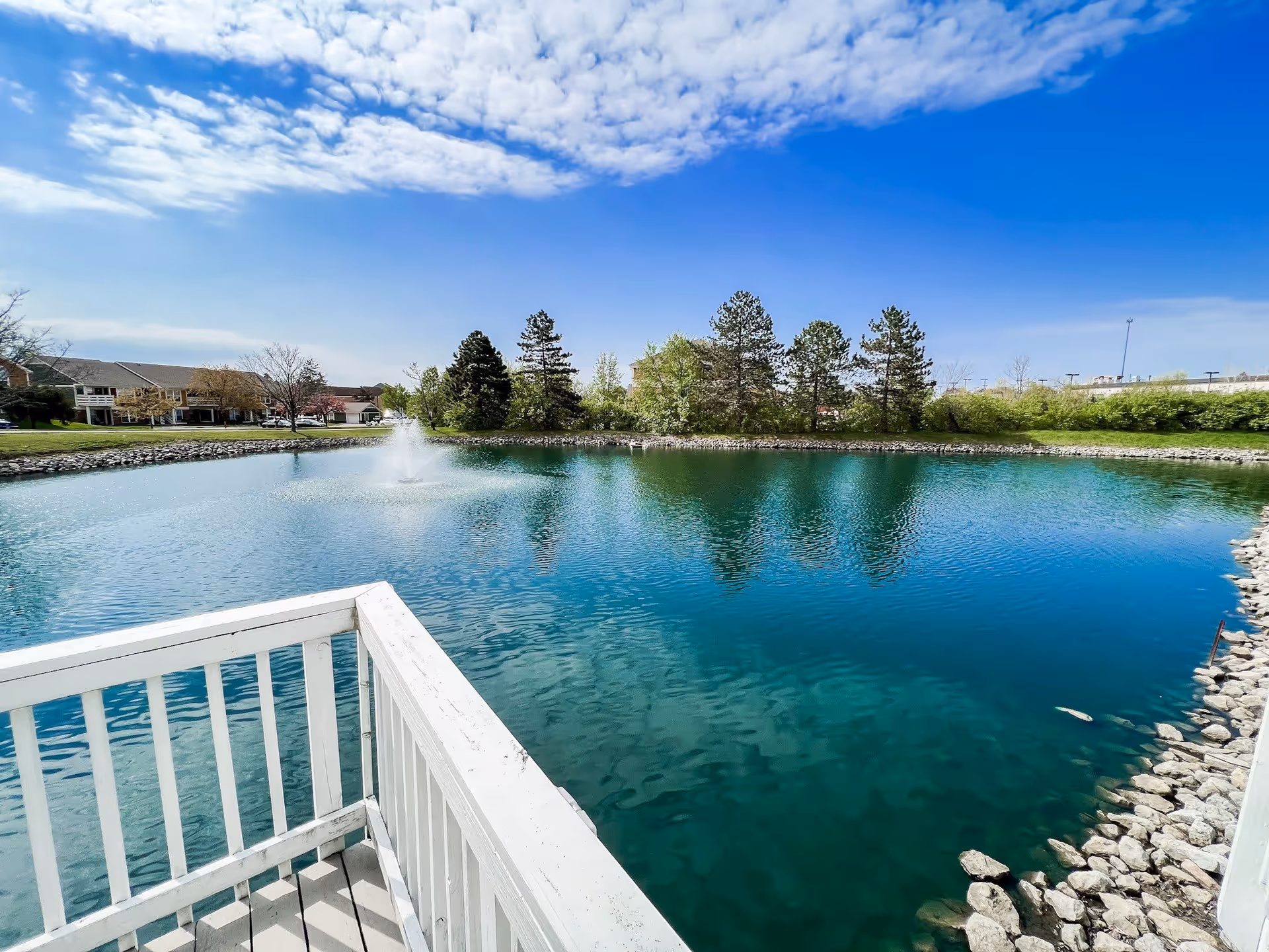 View of a serene pond with a water fountain in the center, surrounded by trees and greenery under a partly cloudy blue sky. A white wooden railing is visible in the foreground, suggesting a deck or balcony overlooking the pond.