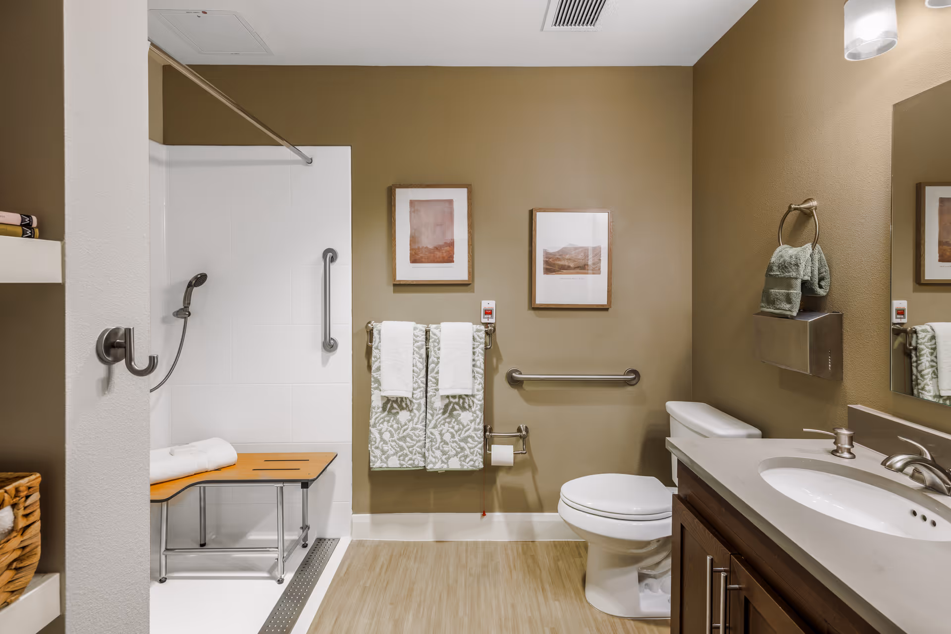 Accessible bathroom featuring a walk-in shower with bench and grab bars, a toilet, sink vanity, and hanging towels.