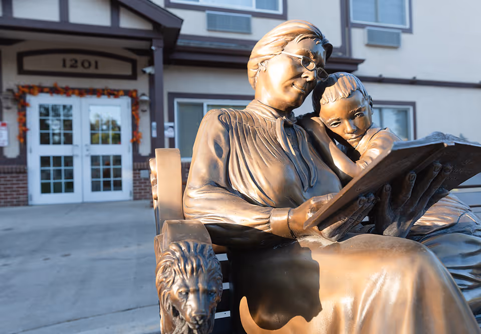 Bronze sculpture of an elder woman reading a book with a child on her lap outside the entrance of an assisted living building.