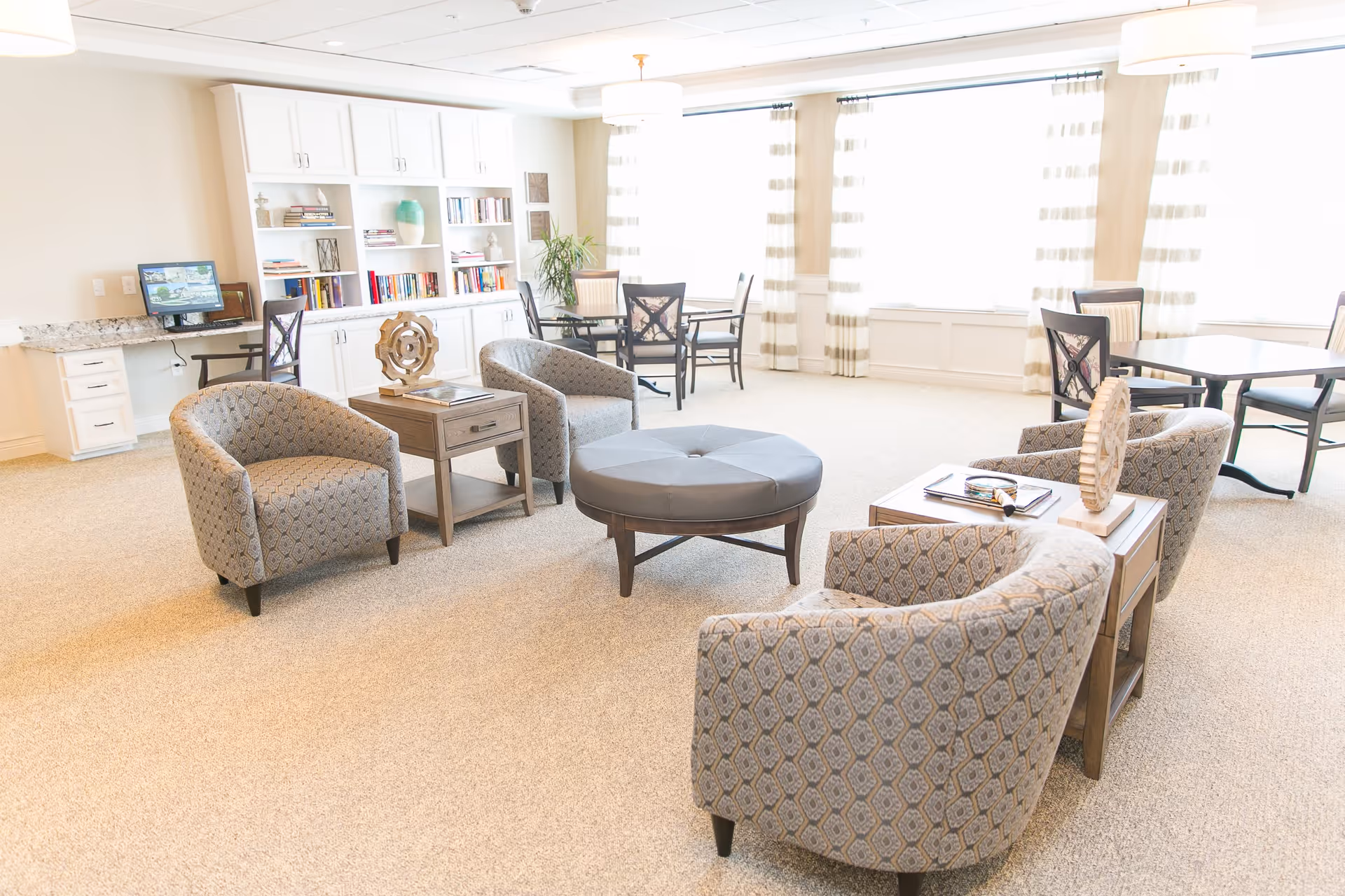 A bright and spacious senior living common area with patterned armchairs arranged around a round ottoman, wooden side tables with decorative items, a built-in white bookshelf filled with books and decor, and several tables with chairs near large windows with sheer curtains allowing natural light to fill the room.