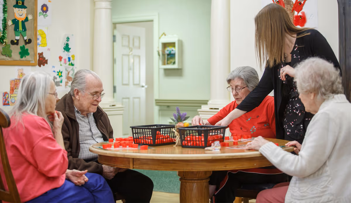 Four elderly individuals sitting around a wooden round table playing a game with red pieces, assisted by a younger woman standing beside them in a well-lit room decorated with colorful artwork on the walls.