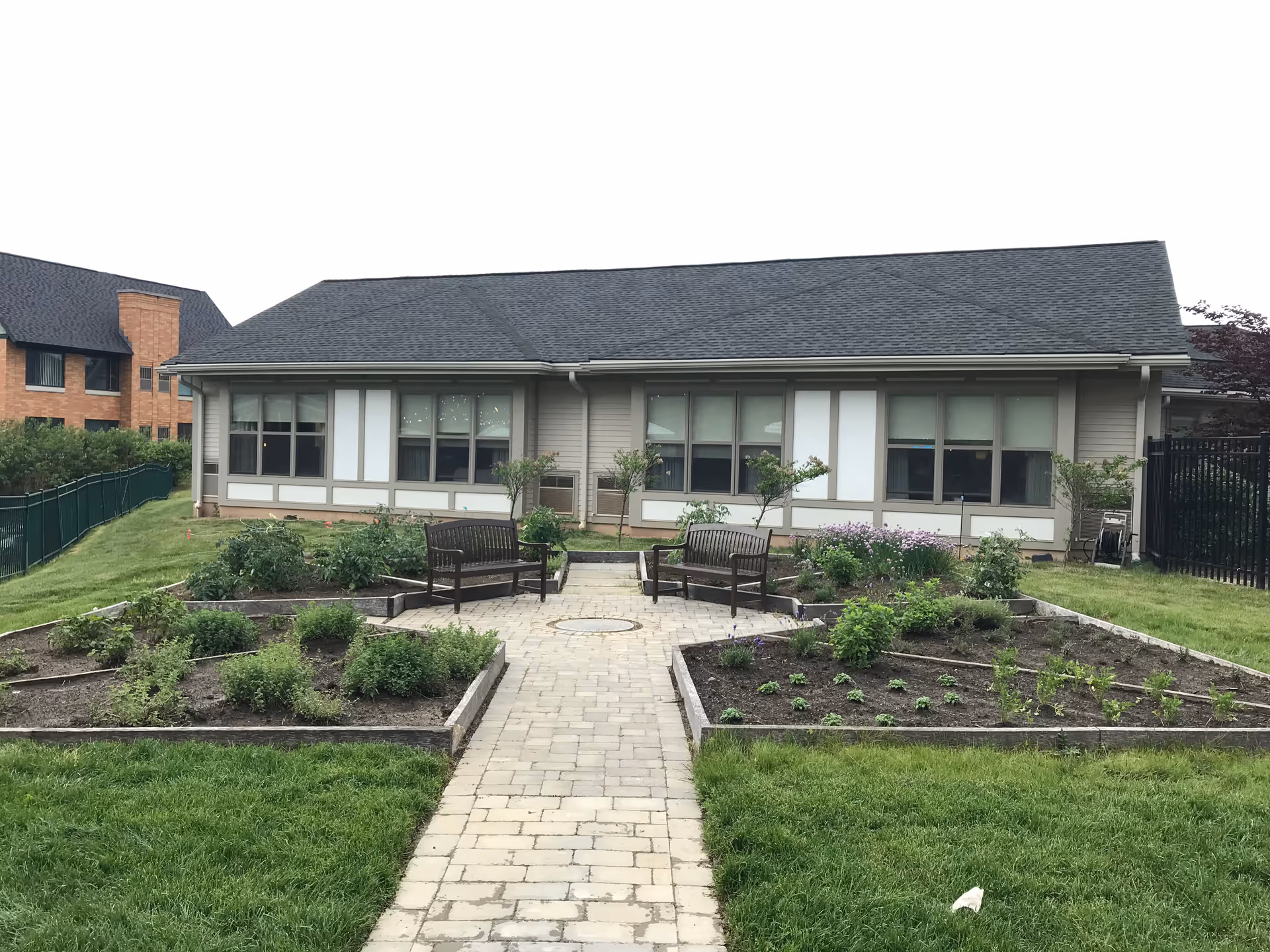 Single-story memory care building with a paved walkway leading to benches and raised garden beds in the foreground.