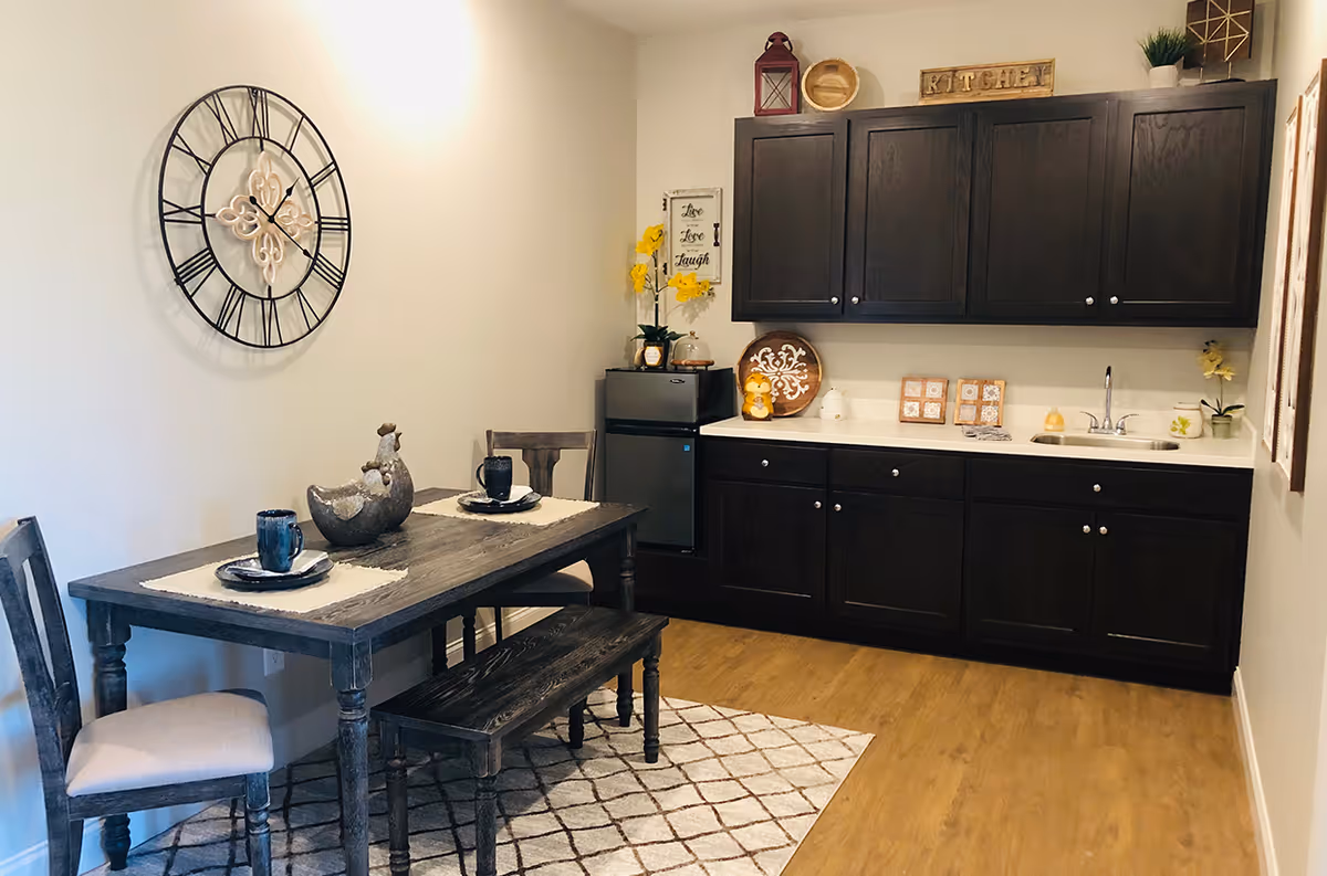 Small dining area with a rustic wooden table, bench and chairs in front of a compact kitchenette with dark cabinets, a mini-fridge and sink.