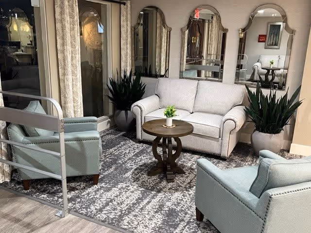 A cozy seating area in a senior living facility featuring a light gray loveseat, two light blue armchairs, a round wooden coffee table with a small plant on it, two large potted plants, and three decorative mirrors on the wall behind the loveseat. The floor is covered with a patterned area rug and there are curtains and glass doors on one side.