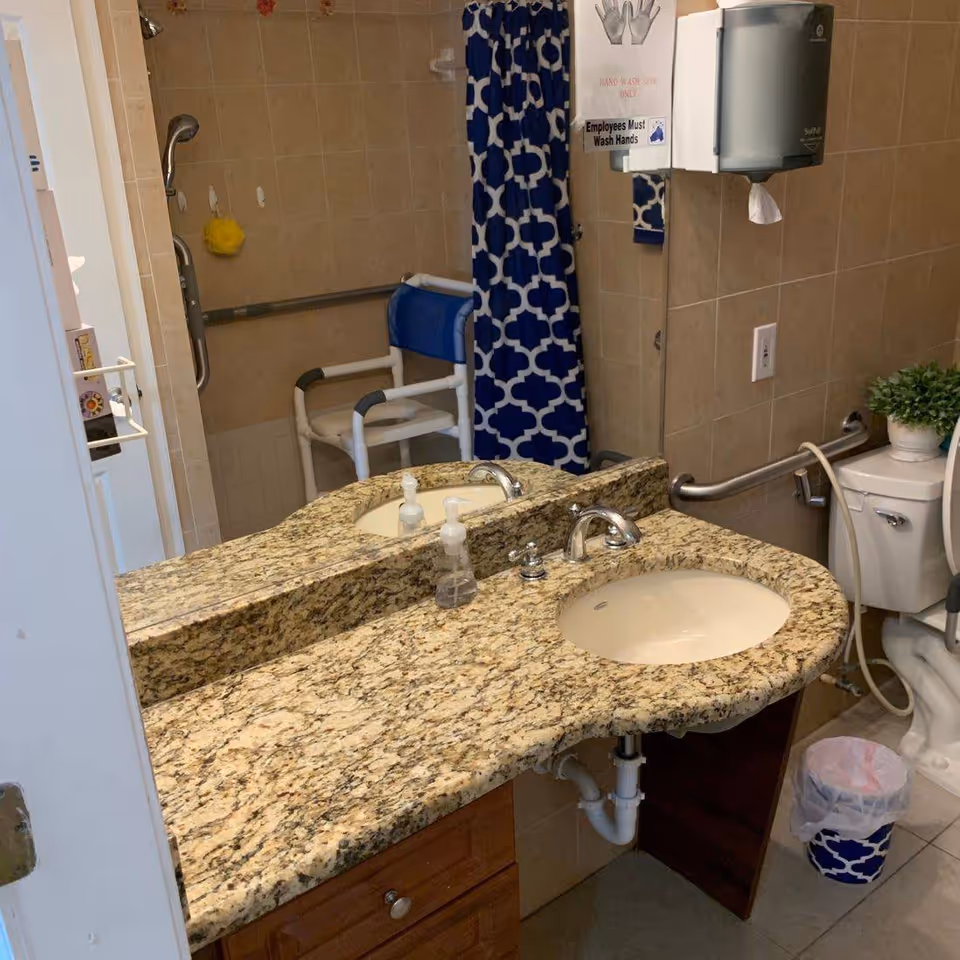 A bathroom with a granite countertop featuring two sinks and chrome faucets. There is a large mirror above the countertop. In the background, there is a shower area with a blue and white patterned shower curtain and a white shower chair. The bathroom also includes a toilet with a grab bar and a small potted plant on top. A soap dispenser and a paper towel dispenser are mounted on the wall, along with a sign that says 'Employees Must Wash Hands.' A small trash can with a blue and white patterned liner is on the floor.