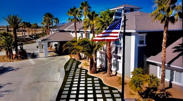 Front exterior of a senior living facility showing a covered driveway entrance, an American flag, and palm trees.