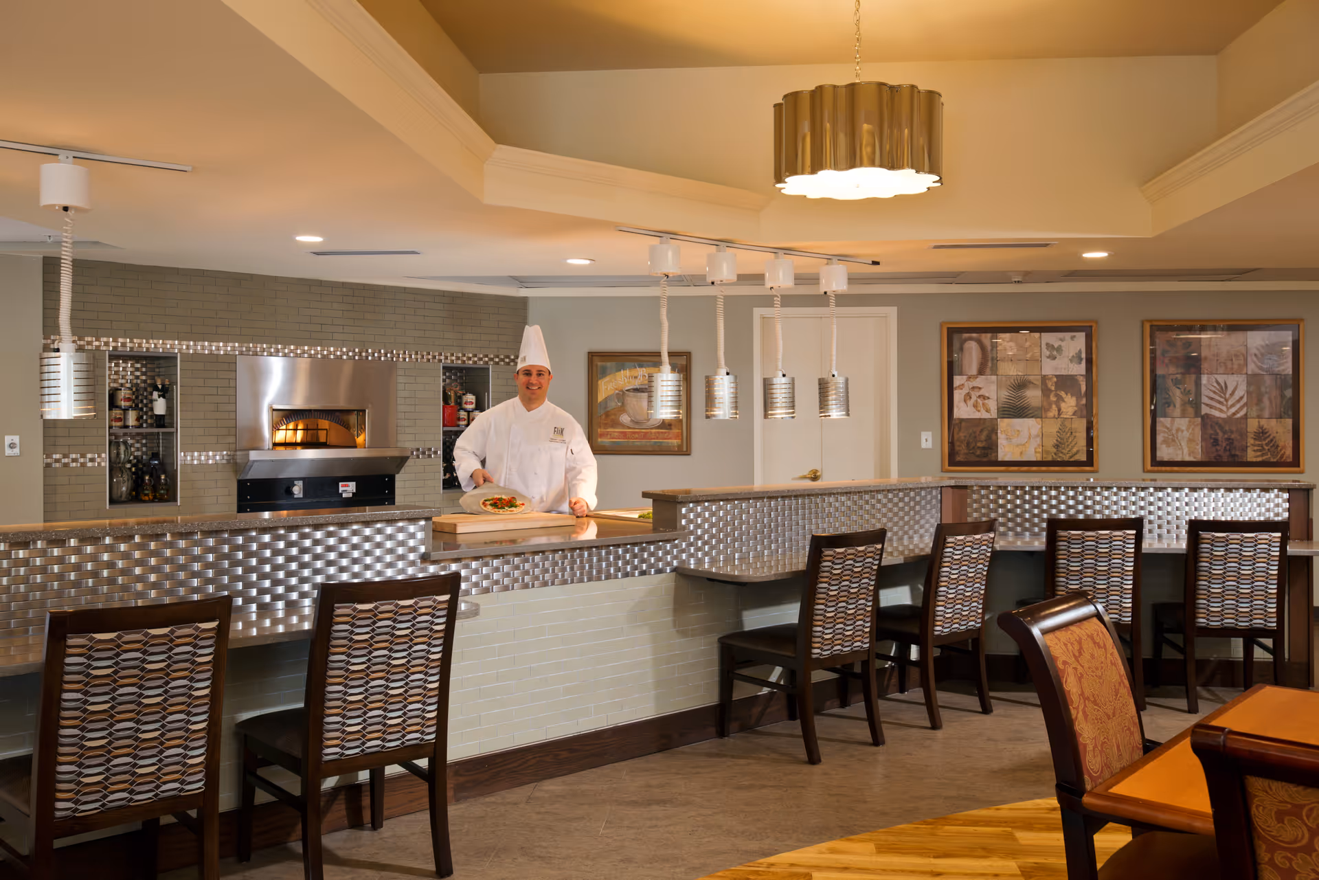 A chef stands behind a tiled serving counter in a dining area with bar seating and pendant lights.