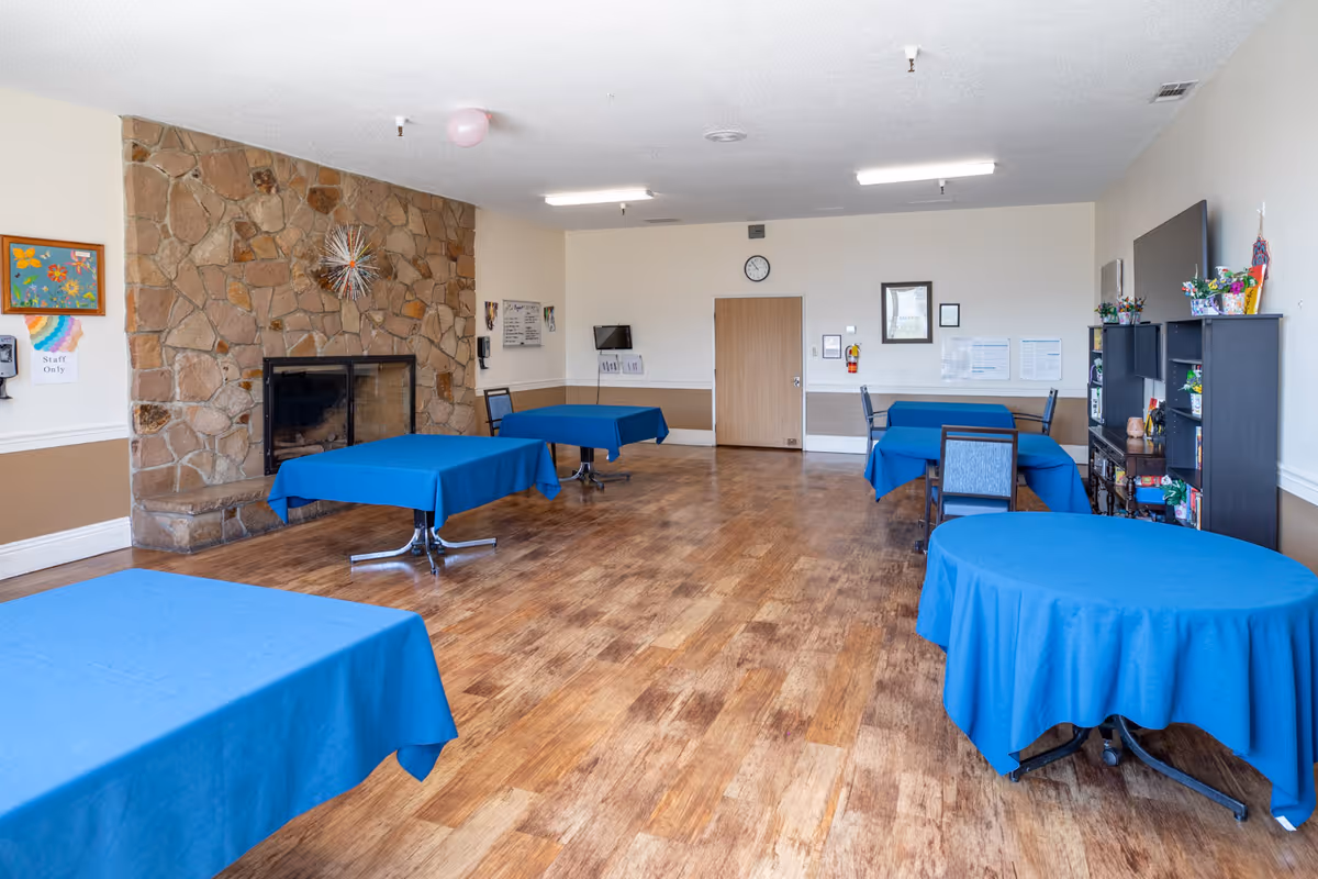 A spacious room with wooden flooring and several tables covered with blue tablecloths. There is a stone fireplace on the left wall, a clock and framed pictures on the far wall, and a shelving unit with decorations and a TV on the right. The room appears clean and well-lit with ceiling lights.