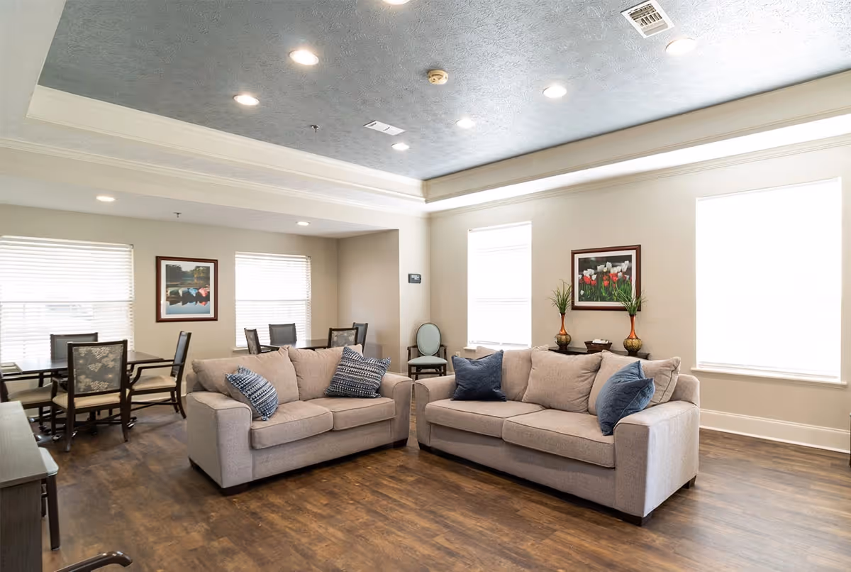 Bright communal living room with two beige sofas, decorative pillows, a dining table with chairs, framed artwork, and large windows.