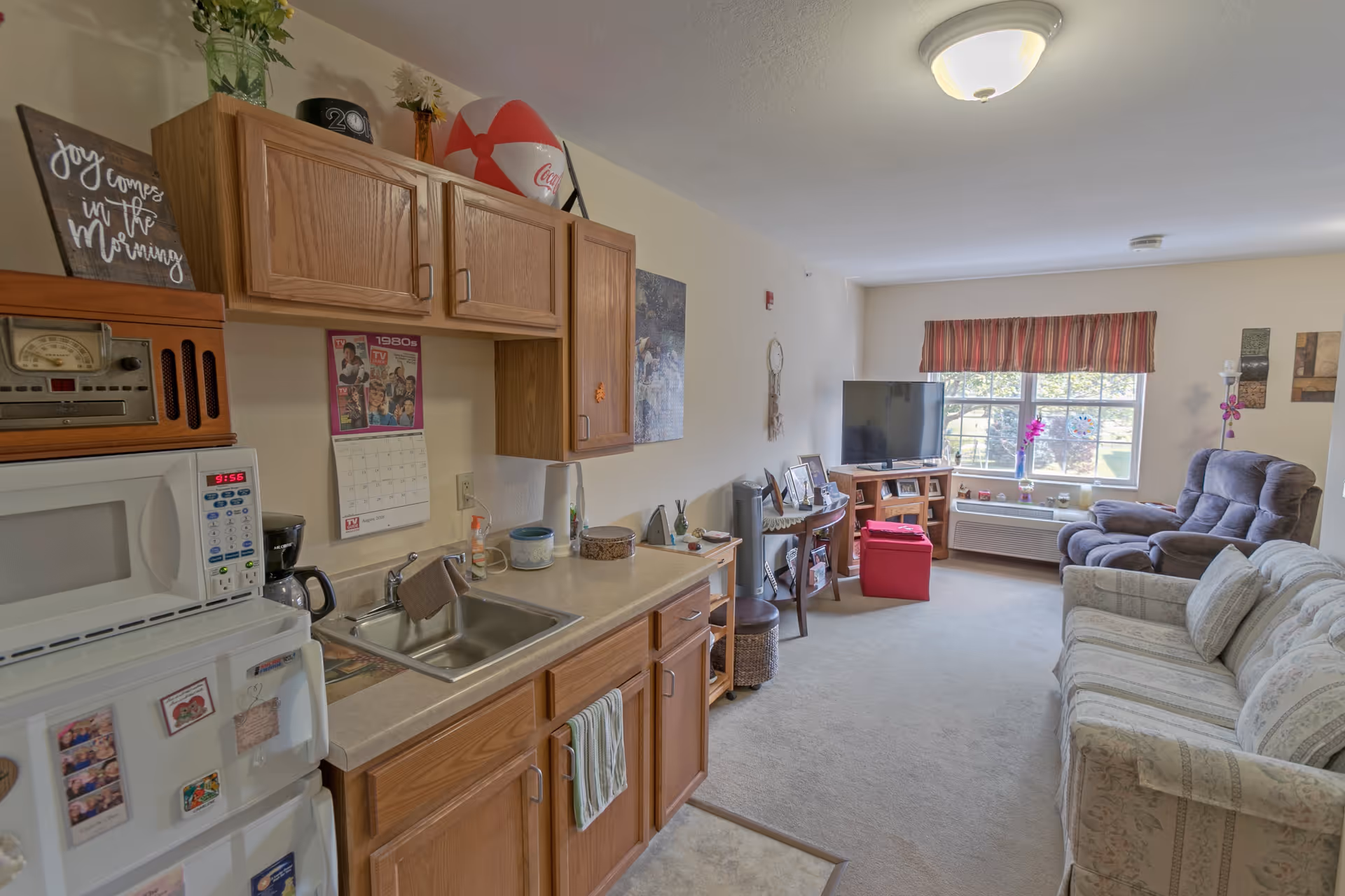 A cozy living area in a senior living facility featuring a small kitchenette with wooden cabinets, a microwave, and a sink on the left. The living room area has a patterned sofa, a recliner chair, a TV on a wooden stand, and a window with a striped valance letting in natural light. Various decorative items and framed photos are placed on surfaces throughout the room.
