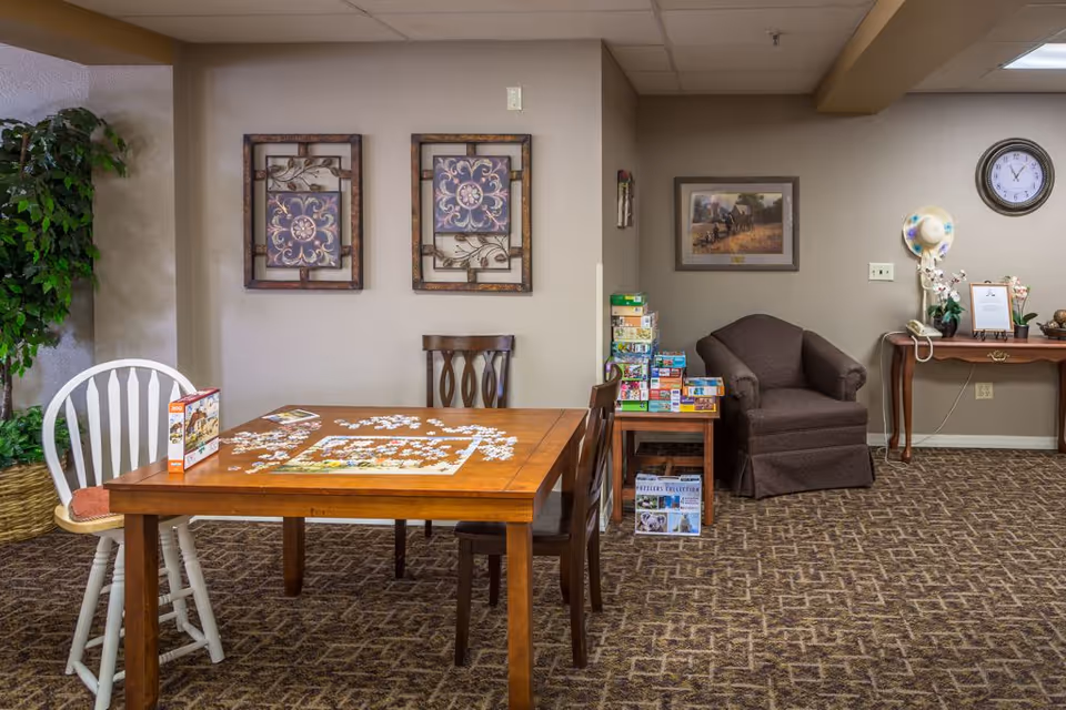 A cozy activity room with a wooden table in the center featuring a partially completed jigsaw puzzle. Two chairs, one white and one wooden, are placed around the table. On the right side, there is a brown armchair next to a small table stacked with various board games and puzzles. The walls are decorated with framed artwork and a clock, and a potted plant is visible on the left side. The carpet has a patterned design.