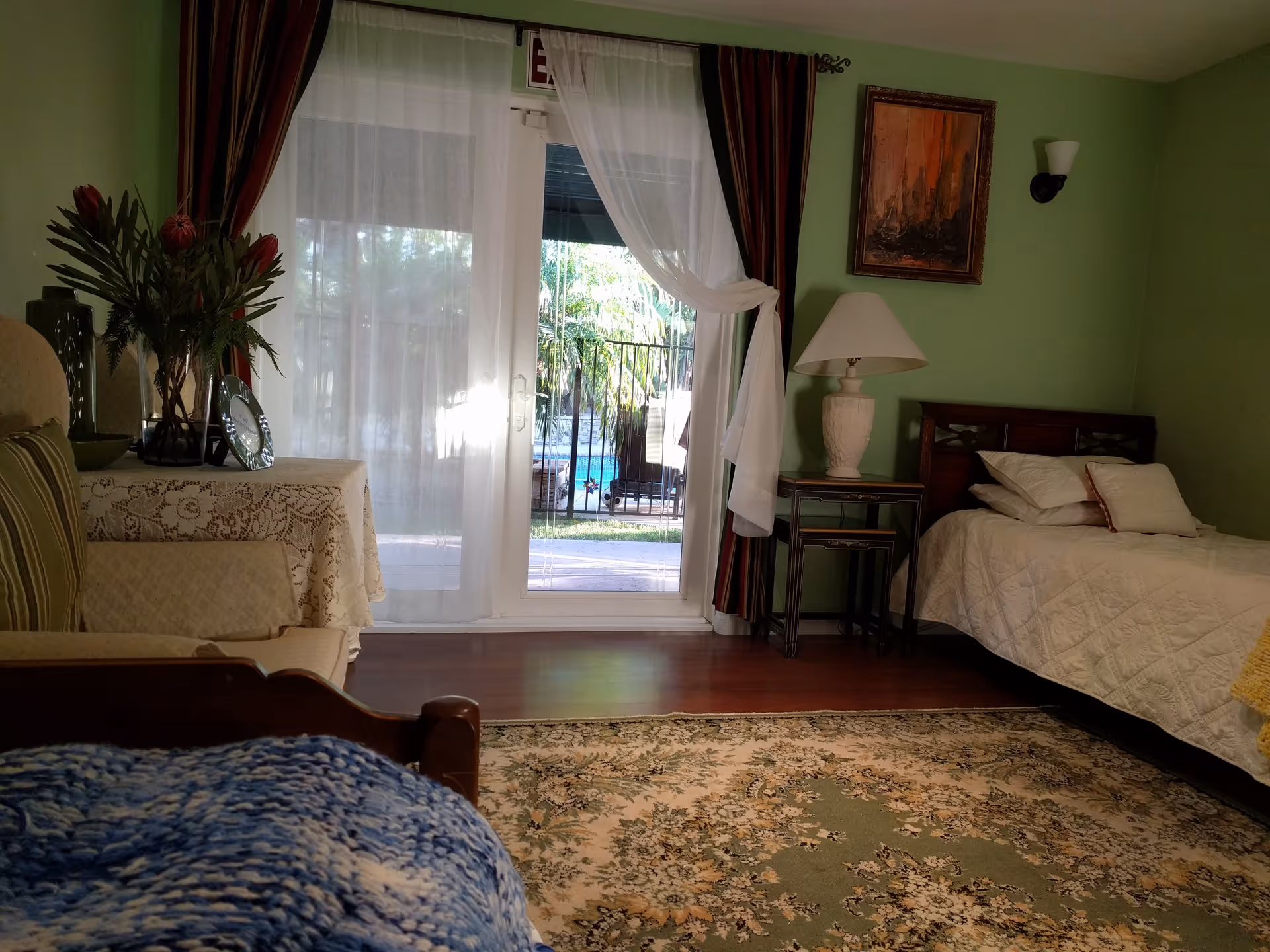 A cozy bedroom with green walls featuring a single bed with white bedding and pillows, a wooden side table with a white lamp, a framed painting on the wall, a table covered with a lace cloth holding a vase with flowers and decorative items, and a glass door with sheer white curtains leading to an outdoor area with a pool and palm trees.