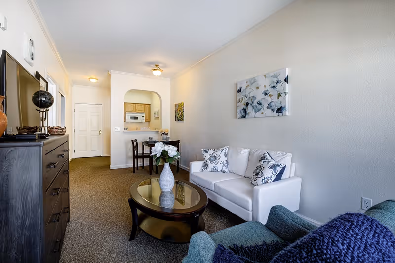 A cozy living room area in a senior living facility with a white couch adorned with patterned pillows, a glass-top coffee table with a white vase of flowers, a green armchair with a blue throw blanket, a dark wooden dresser with a globe and decorative items, and a small dining table with two chairs near a pass-through window to the kitchen.