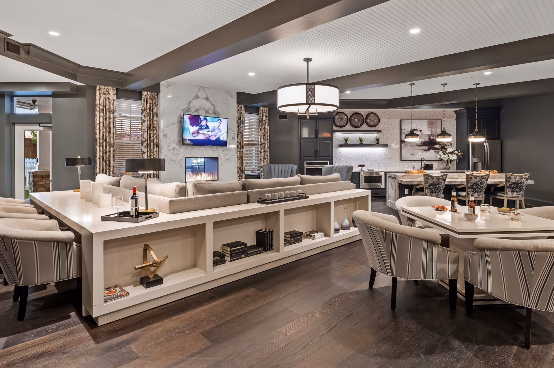 A modern and spacious senior living common area featuring a large beige sectional sofa with built-in shelving, a fireplace with a mounted TV above it, a dining table with six upholstered chairs, and a kitchen area with a marble island, bar stools, and pendant lighting. The room has dark wood flooring, patterned curtains, and decorative elements including books, vases, and artwork.