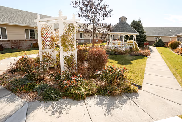 Sunlit courtyard with a white gazebo, trellis, garden beds and paved walkways in front of a single-story building.