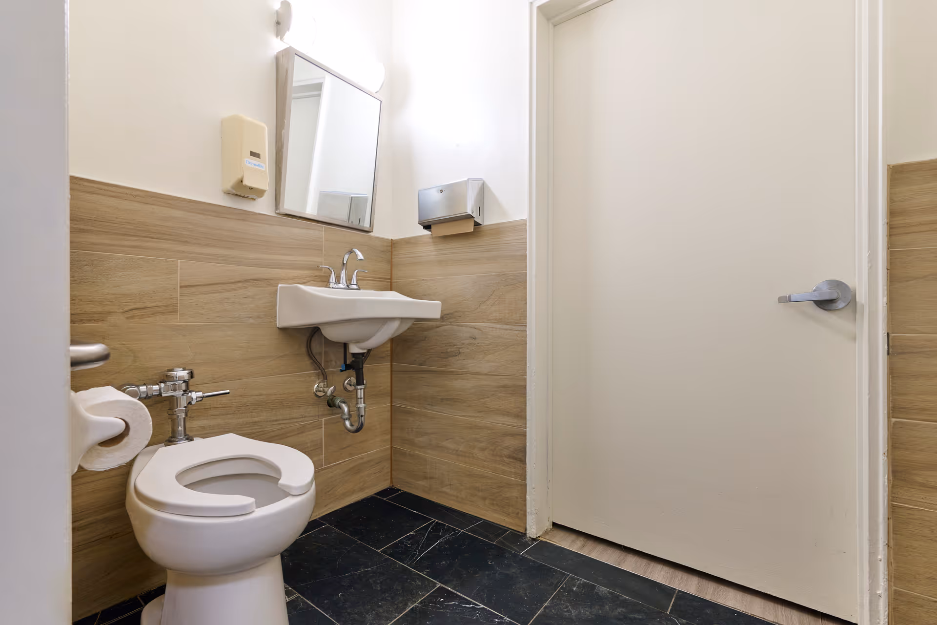 A small bathroom with a white toilet featuring an open front seat, a wall-mounted white sink with a silver faucet, a rectangular mirror above the sink, a beige soap dispenser on the wall, and a silver paper towel dispenser. The walls have light wood paneling halfway up and white paint above, with a black tiled floor.