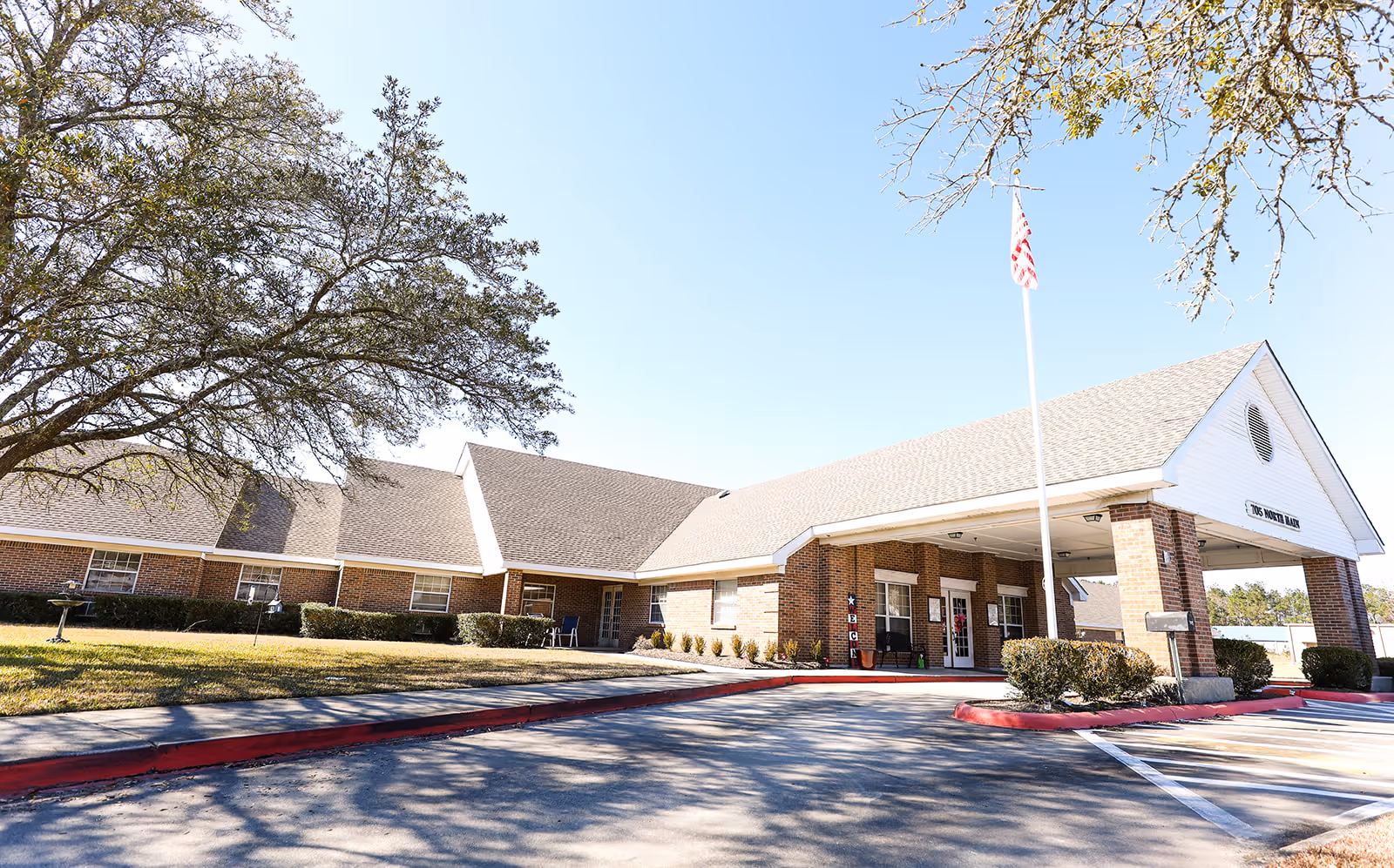 Front entrance of a single-story brick senior living facility with a covered porte-cochere, flagpole, and landscaped lawn.