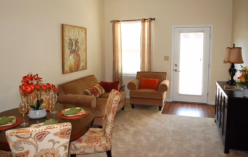A cozy living room with a beige sofa and armchair, both adorned with orange and patterned throw pillows. A round dining table with floral upholstered chairs is set with plates, napkins, and wine glasses. There is a painting of an owl on the wall above the sofa, a window with sheer curtains, and a door with a window letting in natural light. A dark wooden sideboard with a lamp and flowers is positioned against the right wall.