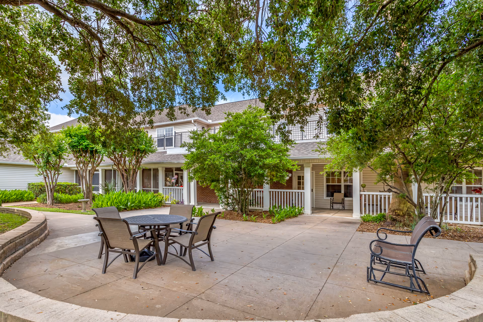 Outdoor patio area at Brookdale Shadowlake with a round metal table and four chairs on a concrete surface, surrounded by trees and greenery. The building in the background has white railings, windows, and a covered porch area.