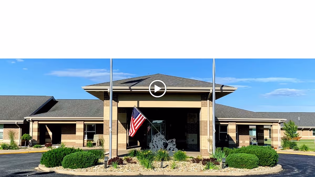 Front entrance of a single-story brick senior living building with a covered porte-cochère, American flag, and landscaped circular driveway.