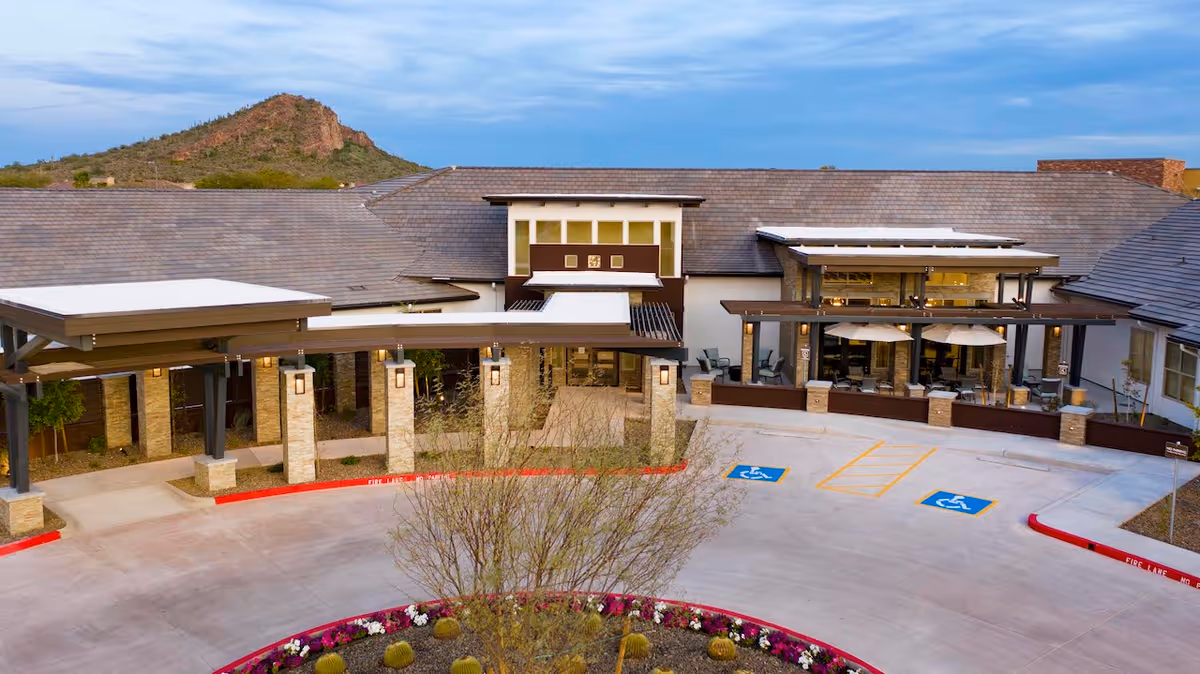 Exterior view of Spring Gardens Senior Living Peoria showing the entrance with covered walkways, handicap parking spaces, and a landscaped circular driveway with flowers and desert plants. A mountain is visible in the background under a partly cloudy sky.