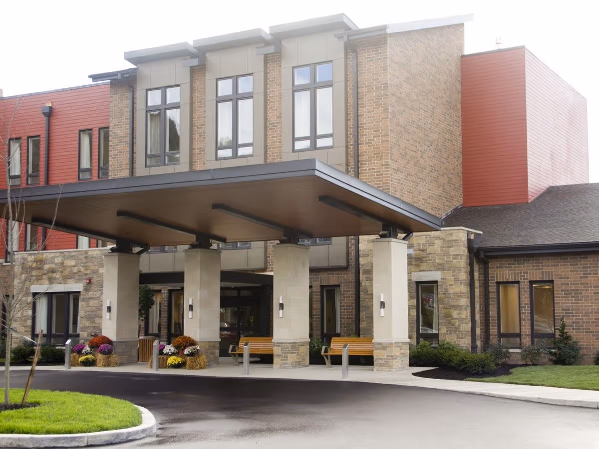 Front exterior view of a modern senior living facility building with a covered entrance supported by four large pillars. There are benches and flower arrangements near the entrance, and the building features a combination of brick and red panel siding with multiple windows.