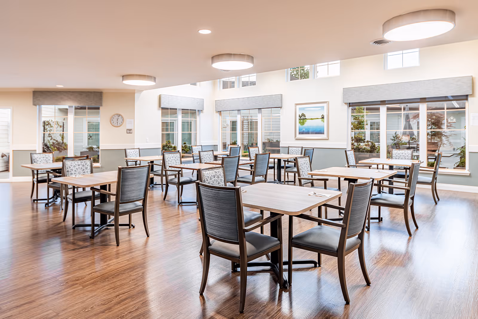 A bright and spacious dining room with multiple wooden tables and cushioned chairs arranged neatly. Large windows with gray valances allow natural light to fill the room, and a framed landscape painting hangs on the wall. The floor is wood, and modern circular ceiling lights illuminate the space.