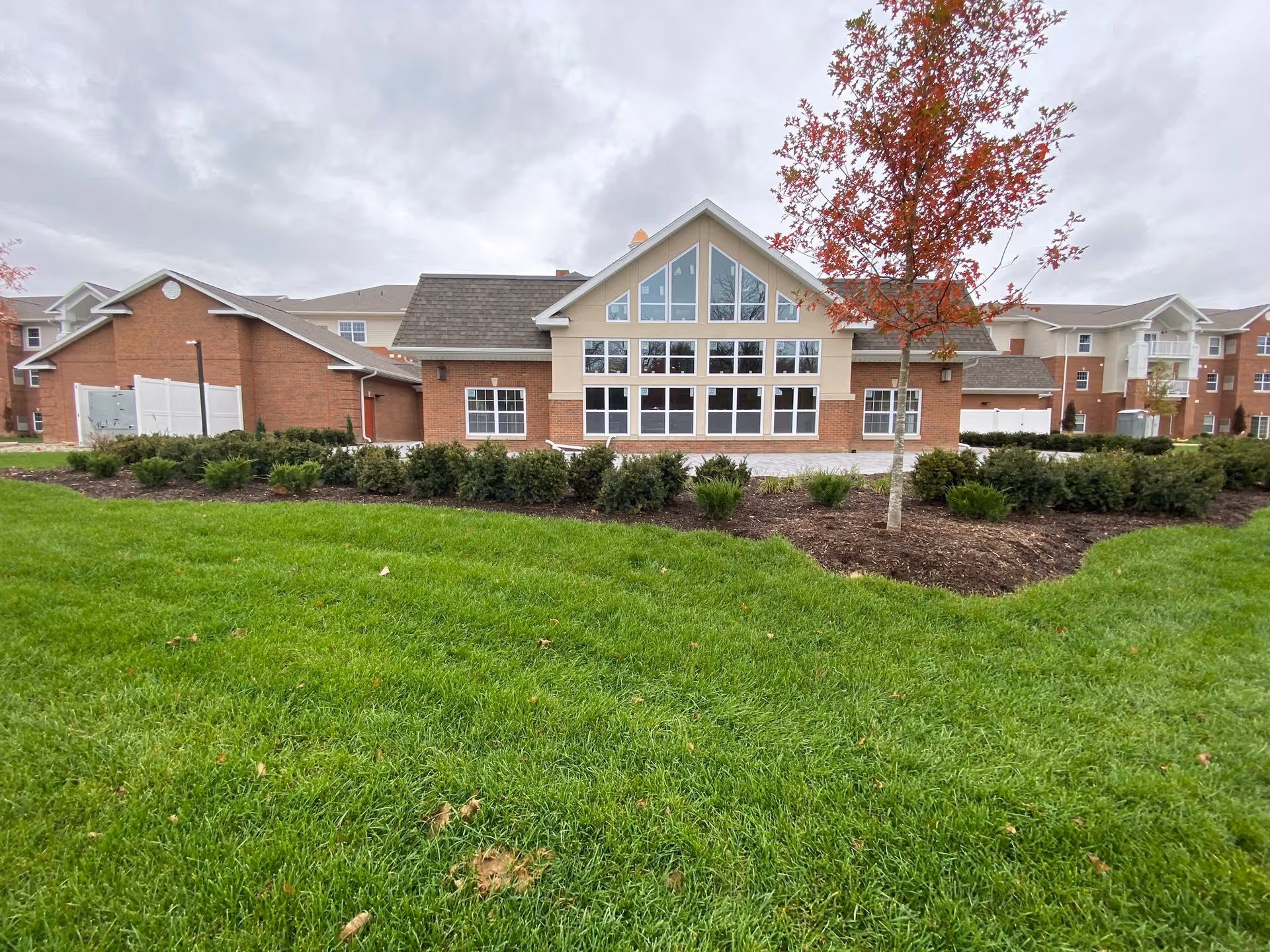 Brick and siding retirement community building with large multi-pane windows, landscaped shrubs and a grassy lawn under a cloudy sky.