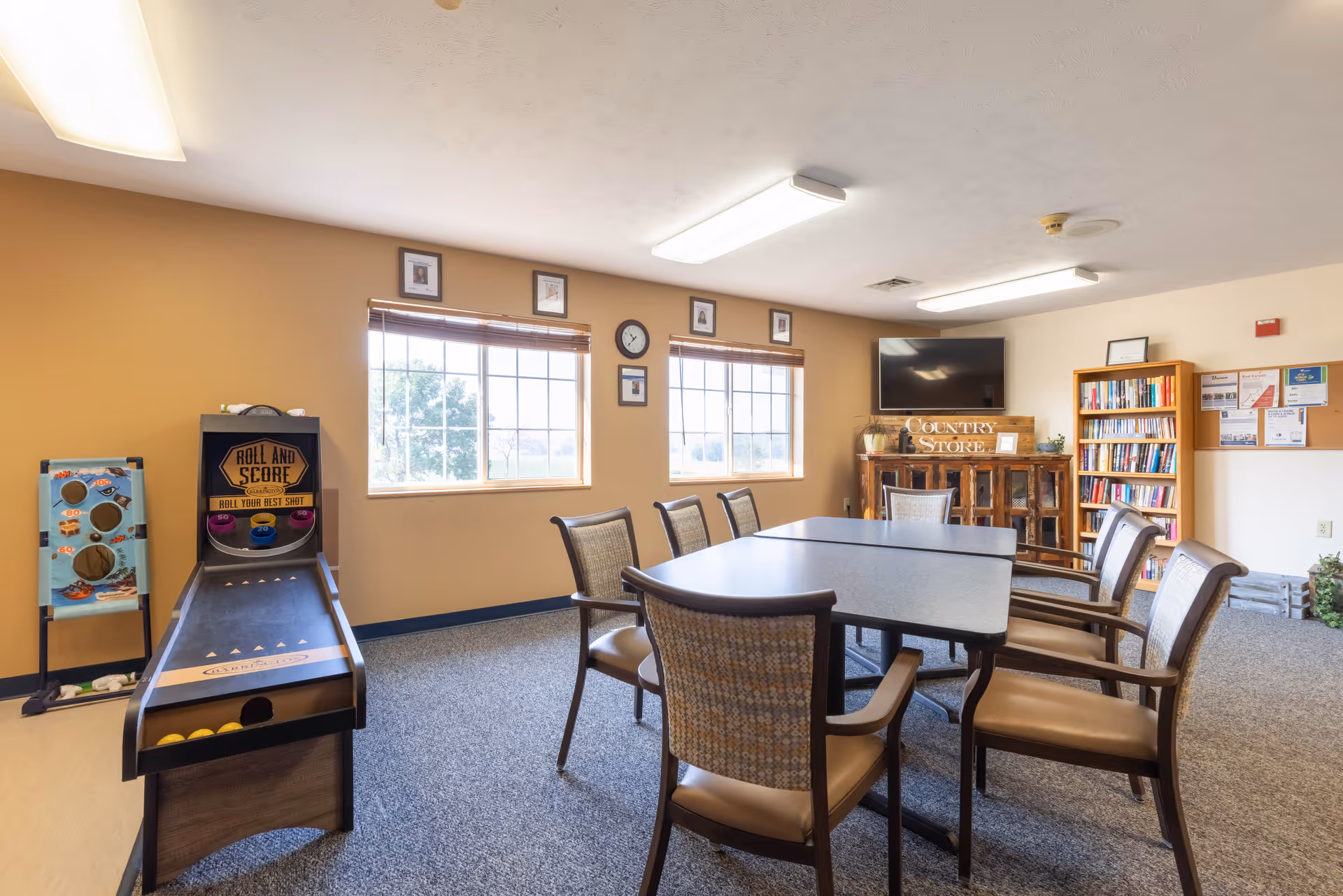A well-lit common room with a rectangular table surrounded by eight chairs. The room features two large windows with blinds, a wall clock, framed pictures, a television mounted on the wall, a wooden cabinet labeled 'Country Store,' a bookshelf filled with books, and two tabletop games including a Roll and Score game and another game with holes for tossing balls.