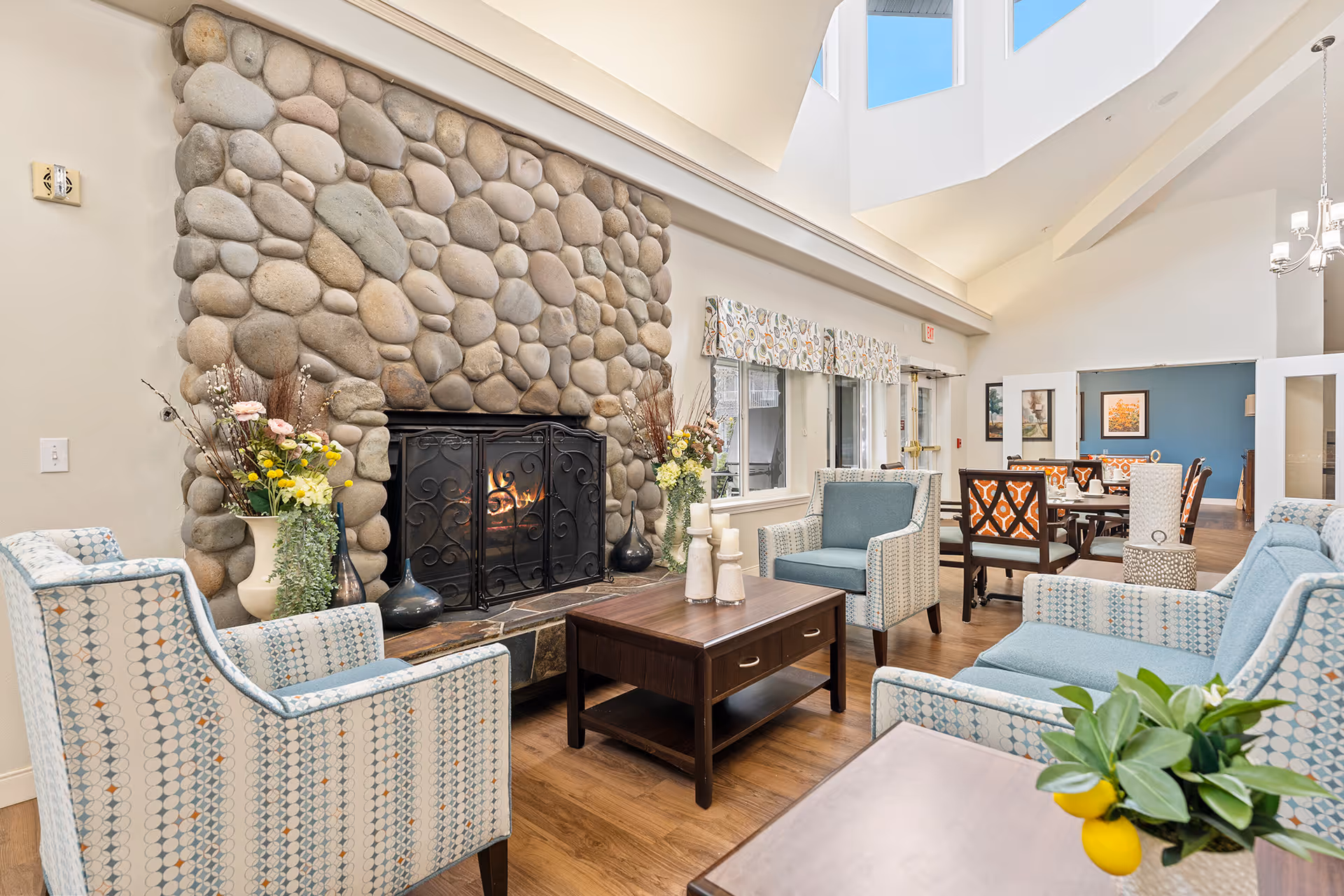 A cozy living room area with a large stone fireplace featuring a fire burning behind a black metal screen. The room has high ceilings with skylights and is furnished with patterned armchairs and a sofa in light blue tones, a dark wooden coffee table with candles, and floral arrangements on either side of the fireplace. In the background, there is a dining area with tables and chairs, and large windows with patterned valances letting in natural light.
