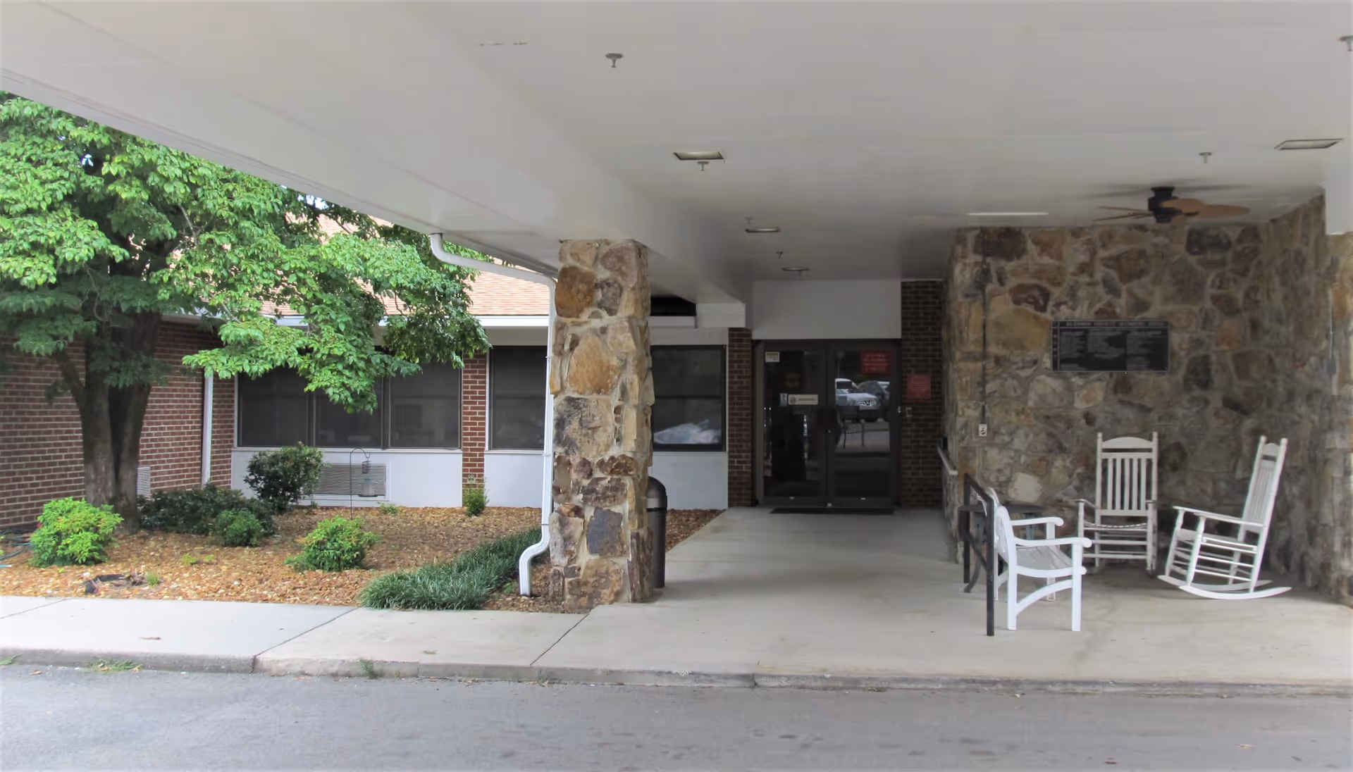 Covered entrance area of a nursing home with stone pillars and walls, three white rocking chairs, a bench, and a ceiling fan. There are bushes and a tree to the left side near the building.