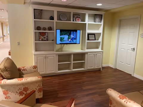 A cozy living room area with floral patterned armchairs and a built-in white entertainment center featuring shelves with decorative items and a flat-screen TV mounted in the center. The room has wooden flooring, soft yellow walls, and a white door on the right side.