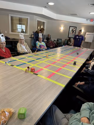 A group of elderly people sitting around a large table with a grid made of yellow and pink tape. Small animal figurines and dice are placed on the table. Some participants are wearing paper crowns. A staff member stands at the far end of the room, smiling. The room has beige walls, windows, and framed artwork.