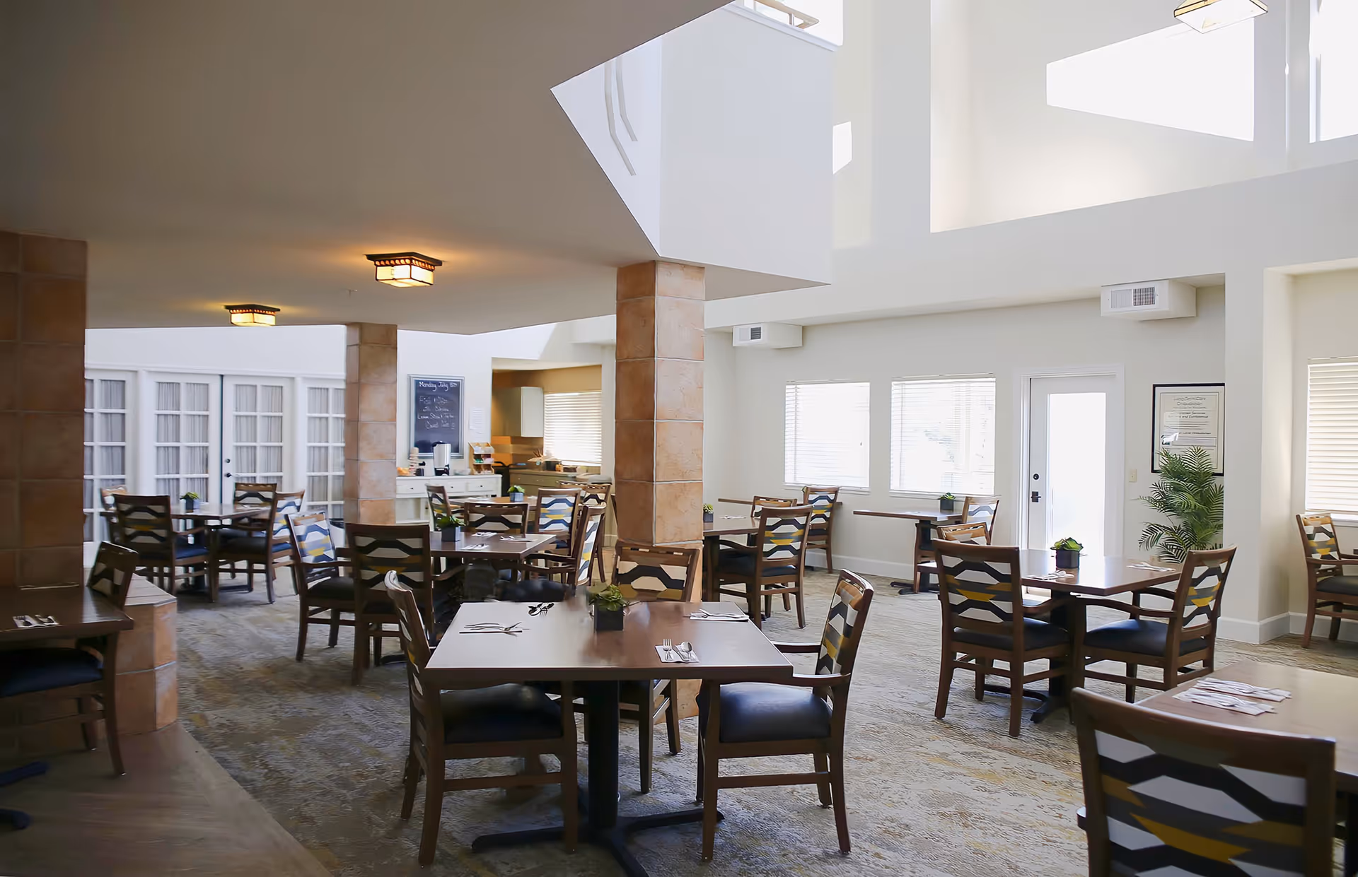 Bright communal dining room with multiple wooden tables and chairs, high ceilings, and a serving area in the background.