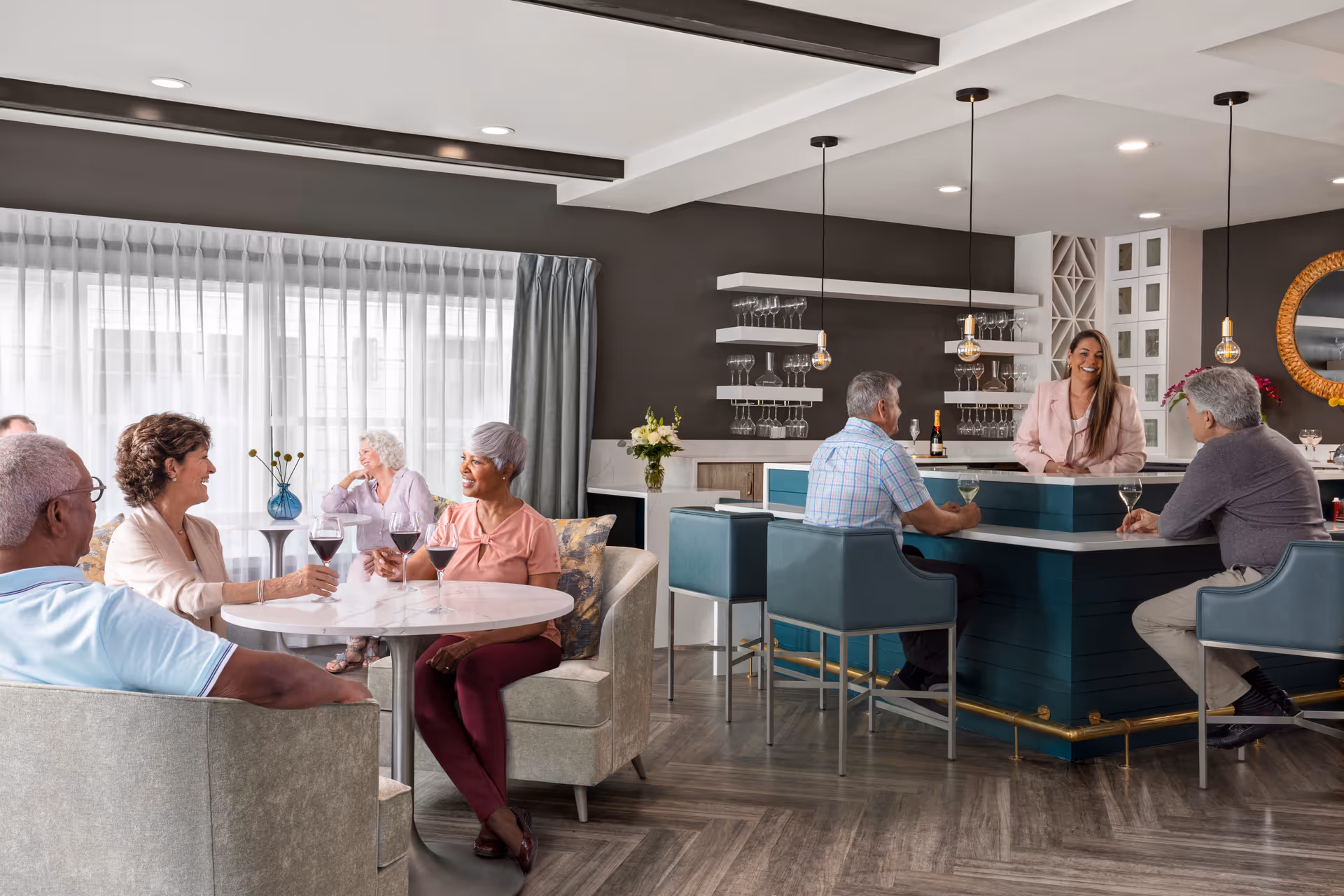 A group of elderly people socializing in a modern lounge area with a bar. Three people are seated around a small round table with wine glasses, while three others are seated at the bar counter where a woman is standing behind it. The room has large windows with sheer curtains, contemporary lighting, and shelves with glassware.