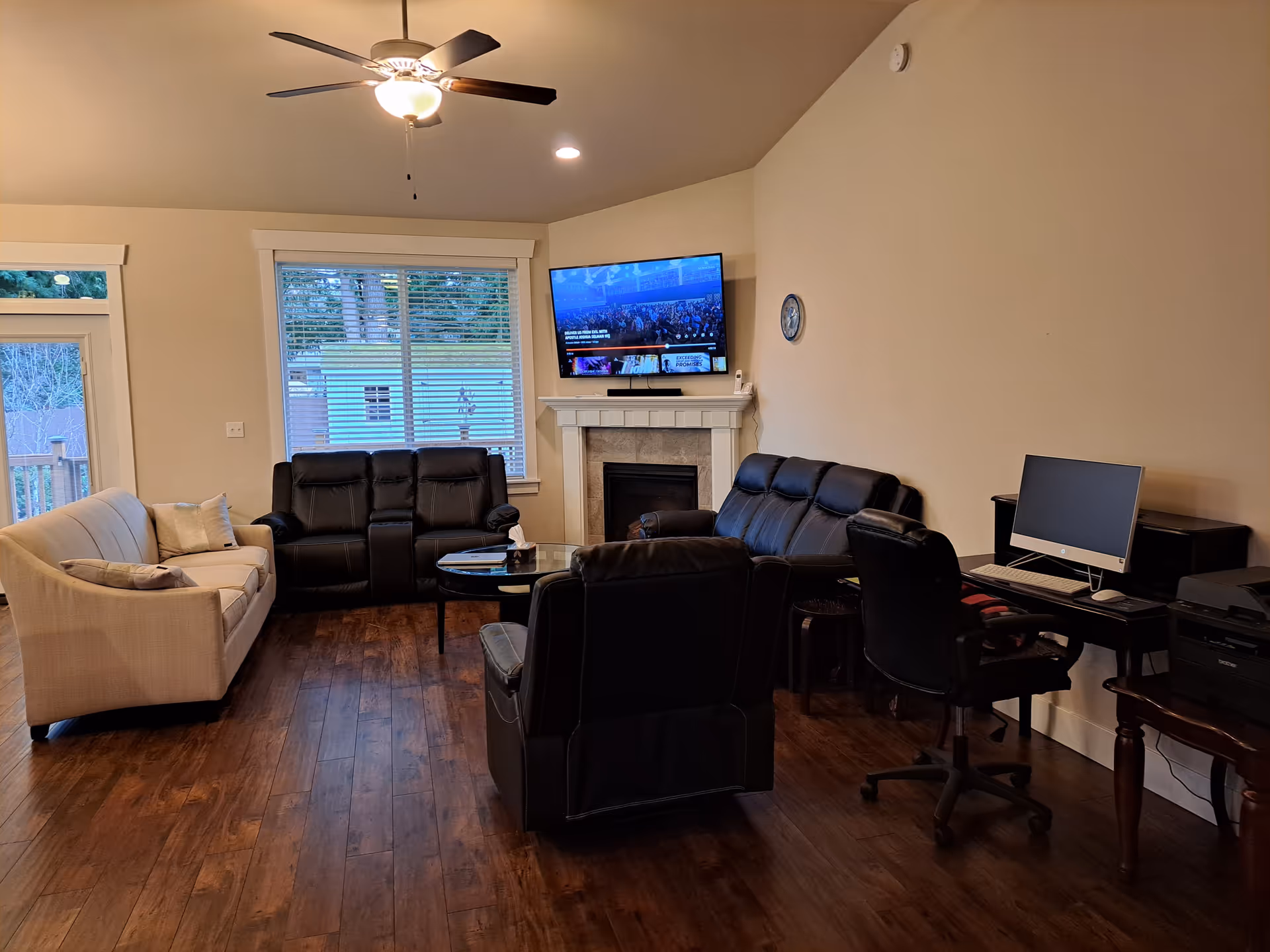 A living room with a ceiling fan, a wall-mounted TV above a fireplace, two black leather sofas, a beige fabric sofa, a glass coffee table, and a desk with a computer and office chair. Large windows and a door provide natural light, and the floor is wooden.
