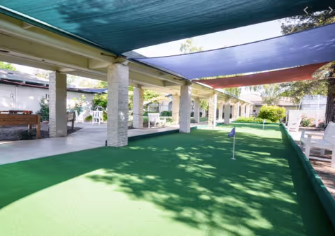 Outdoor putting green area with a small flag, shaded by colorful fabric canopies and supported by stone pillars, with white chairs and tables along the sides.
