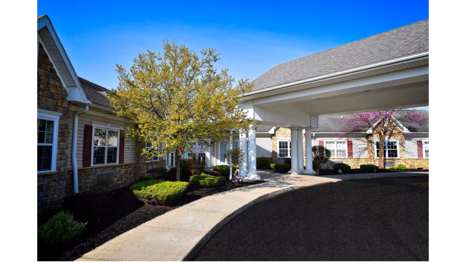 Exterior view of a senior living facility with a covered driveway entrance, stone and siding walls, windows with red shutters, and landscaped greenery including trees and bushes under a clear blue sky.