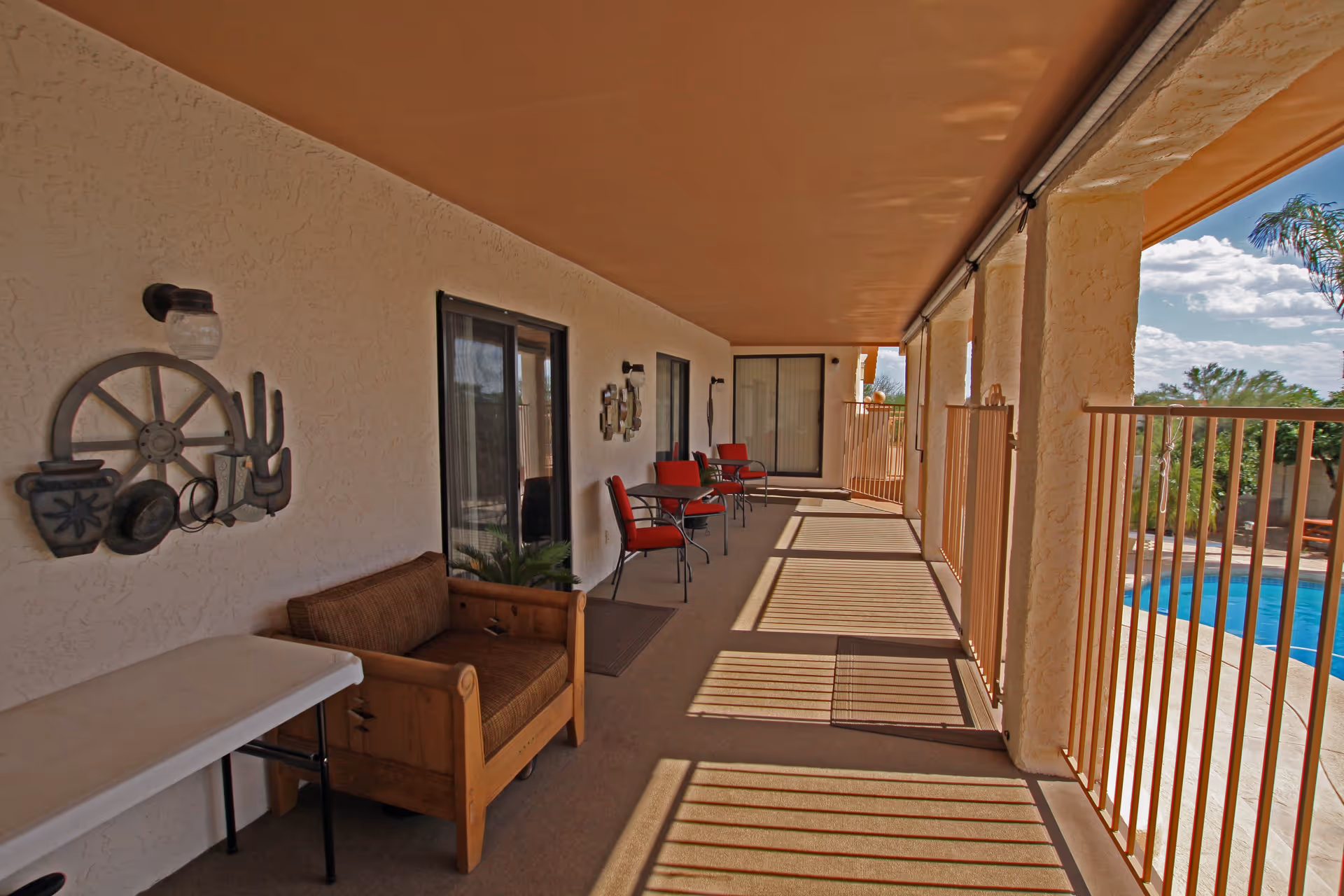 Covered outdoor patio area with a wooden armchair, a folding table, and several red cushioned chairs with small tables. The patio has a textured beige wall with decorative metal art and sliding glass doors leading inside. The patio overlooks a fenced swimming pool and a garden with trees under a partly cloudy sky.