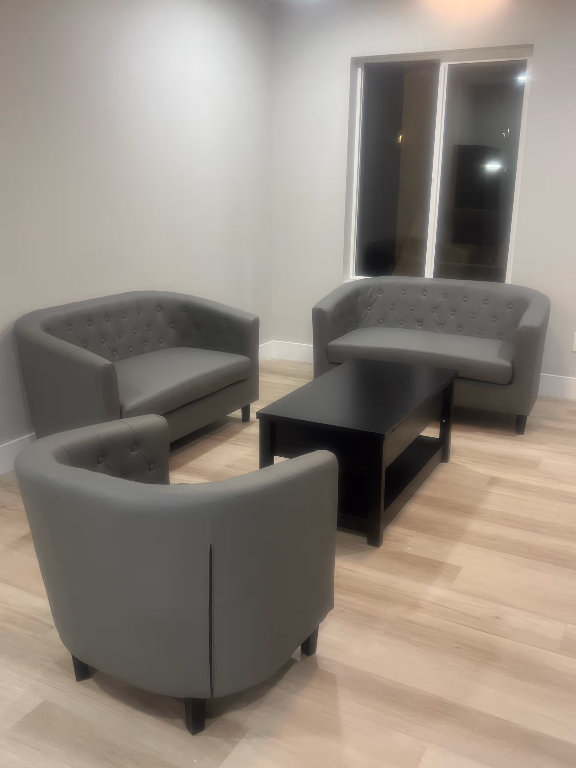 A small seating area with two gray tufted loveseats and one matching armchair arranged around a black rectangular coffee table. The room has light wood flooring, white walls, and a window with a white frame.