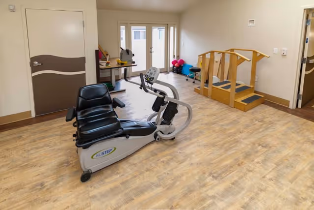 A rehabilitation room with a NuStep exercise machine in the center, a wooden set of stairs with handrails on the right, a table with therapy equipment in the back left corner, and double glass doors letting in natural light.