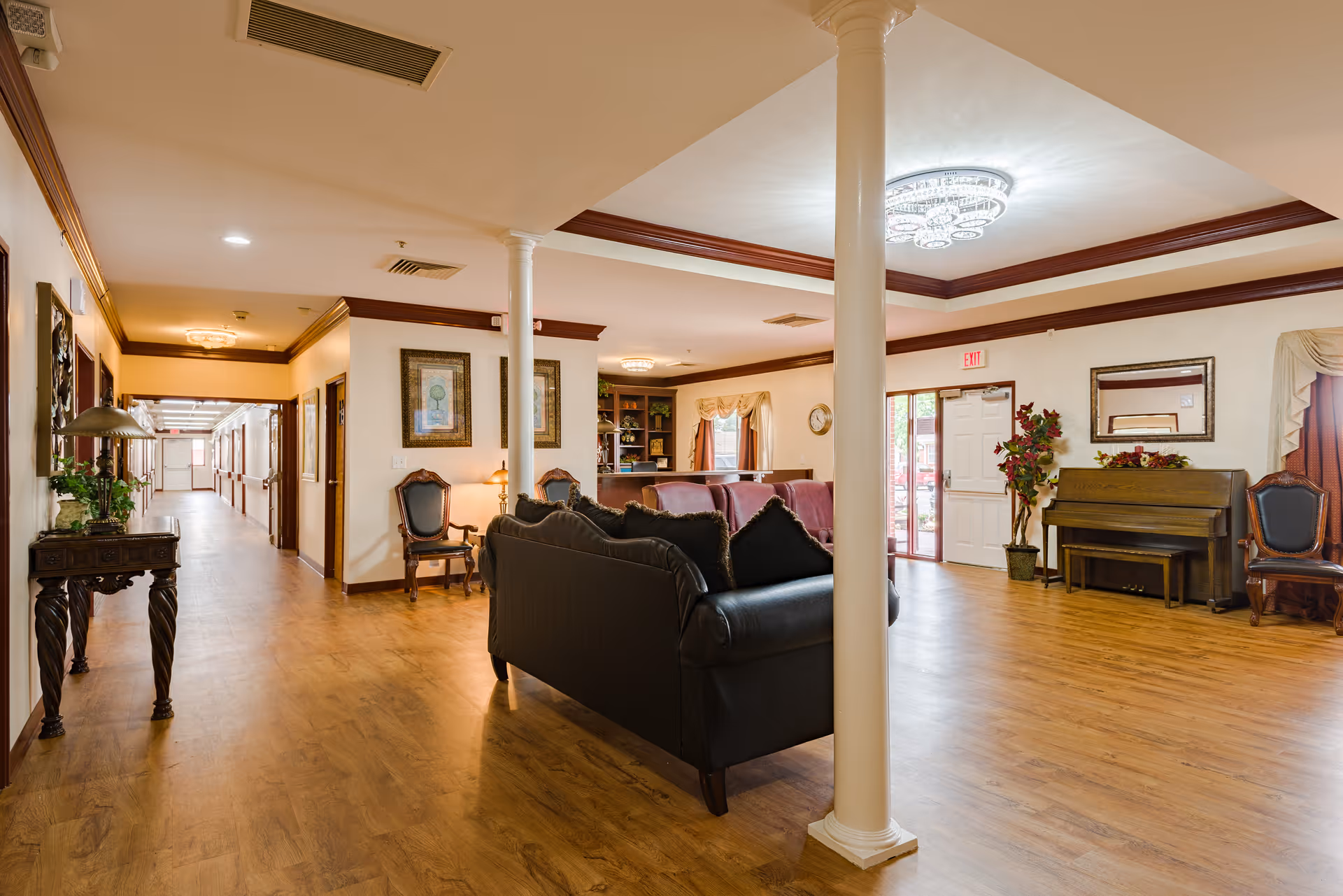 Well-lit common lounge in a retirement community with sofas, chairs, a piano, decorative columns and a hallway.