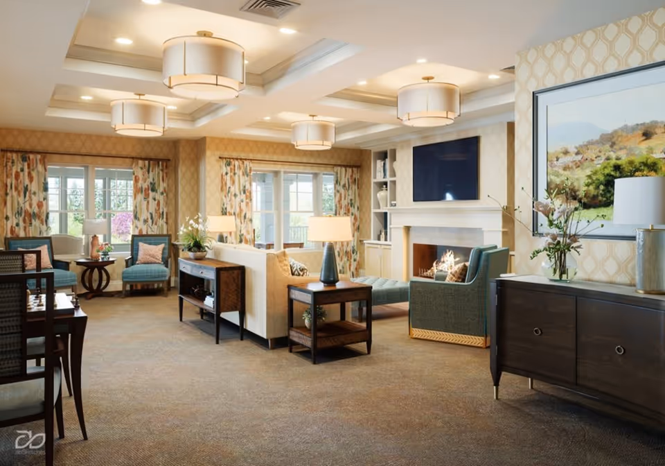 A spacious and well-lit living room with beige walls and carpeted floor. The room features a white sofa, blue armchairs, wooden side tables with lamps, a fireplace with a fire burning, a large flat-screen TV mounted above the fireplace, and large windows with floral curtains letting in natural light. There is also a dark wooden cabinet with a vase of flowers and a large framed landscape painting above it.