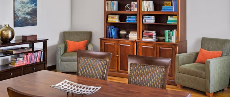 A cozy interior room featuring a wooden dining table with a decorative centerpiece, two patterned chairs, two green armchairs each with an orange pillow, a wooden bookshelf filled with books and decorative items, and a side table with a gold vase and more books. A piece of abstract art hangs on the wall.