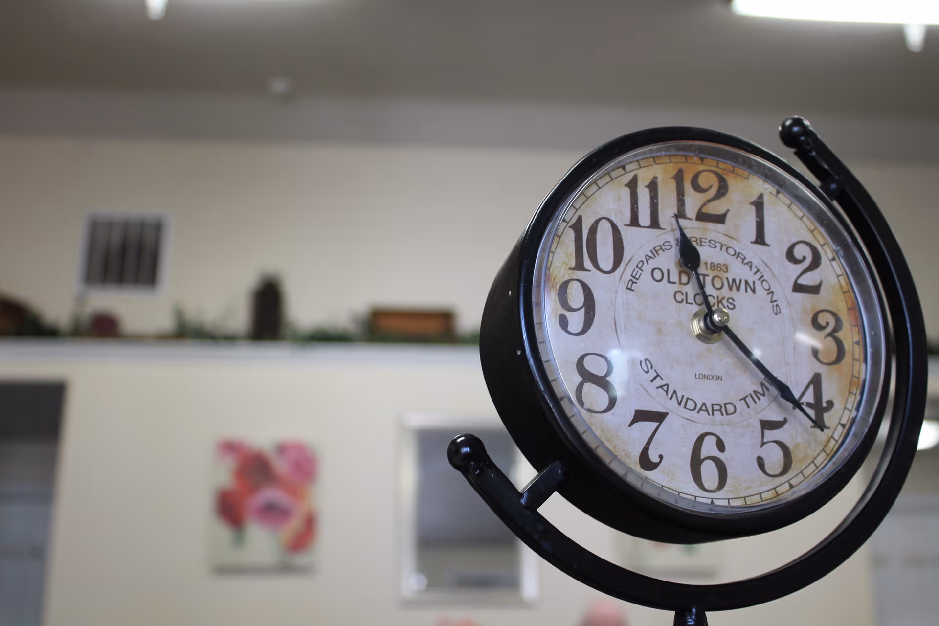 Decorative round vintage-style clock on a stand indoors with a blurred wall and artwork behind it.