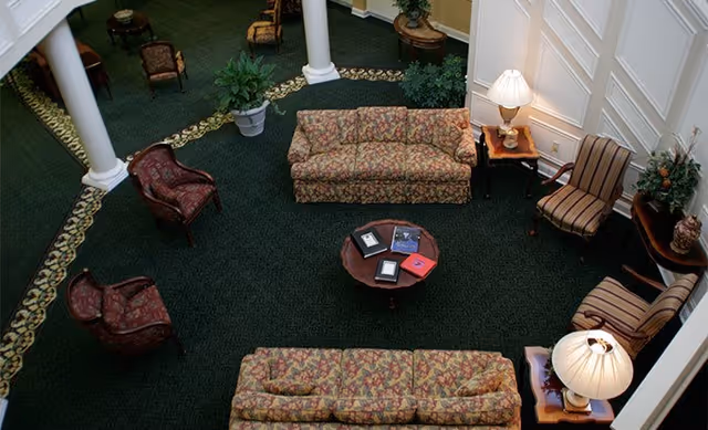 Overhead view of a sitting area in a senior living facility featuring two floral patterned sofas, two striped armchairs, two red patterned armchairs, a round wooden coffee table with books on it, side tables with lamps, potted plants, and white columns on a dark green carpet with a decorative border.