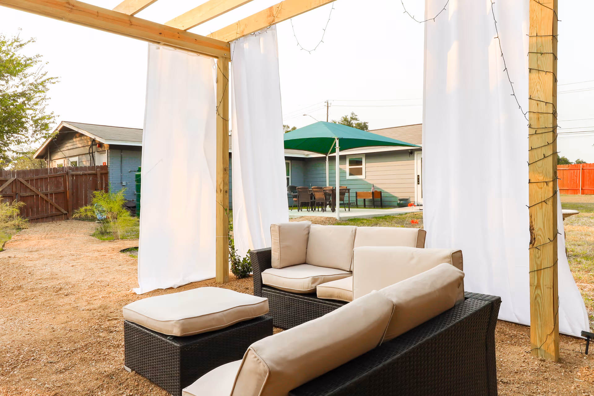 Outdoor seating area with cushioned wicker furniture under a wooden pergola with white curtains and string lights. In the background, there is a green patio umbrella over a table and chairs, a wooden fence, and a small building.
