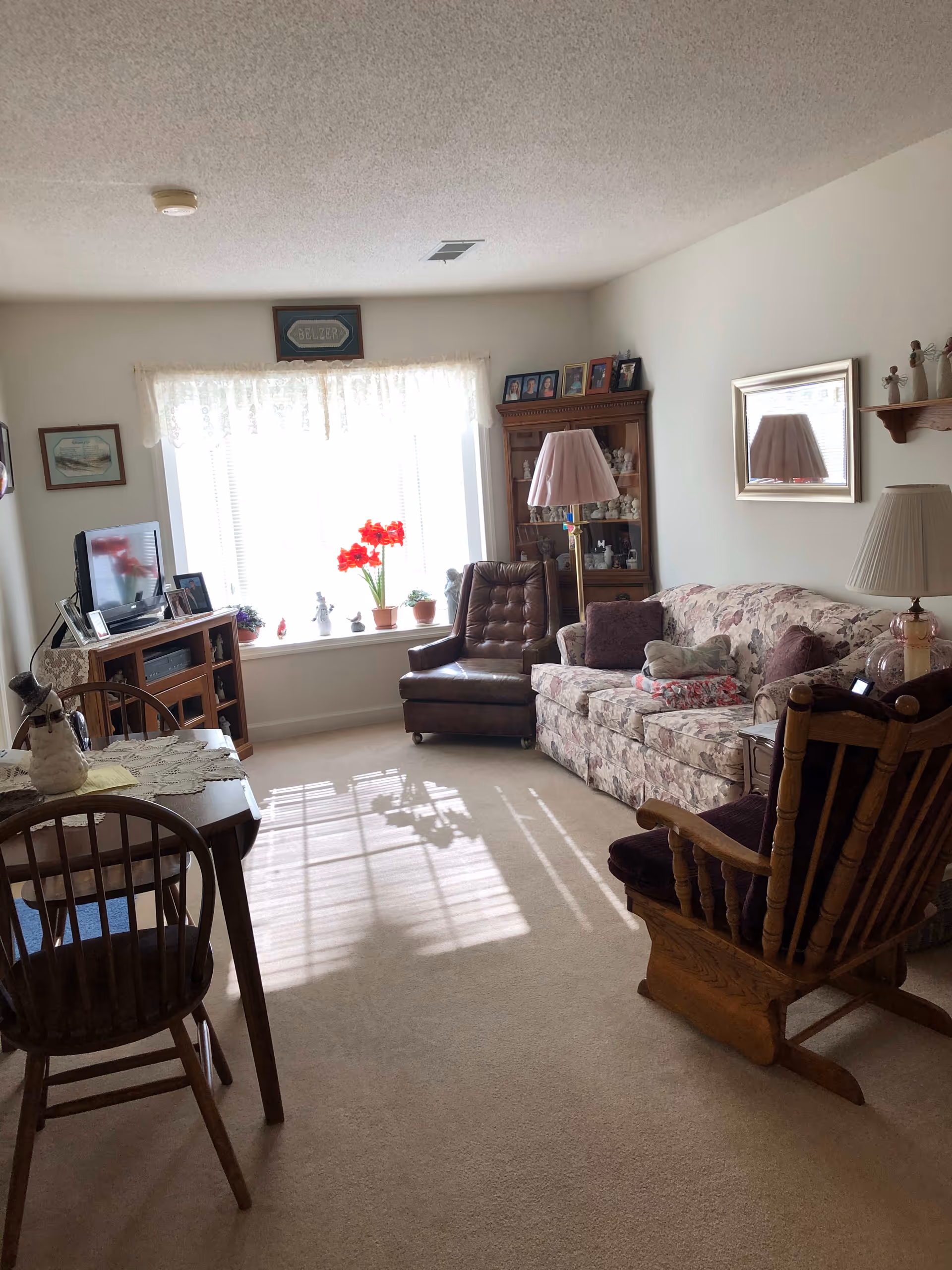 A cozy living room with a floral patterned sofa, a brown leather armchair, and a wooden rocking chair. There is a wooden cabinet with glass doors displaying decorative items, a floor lamp with a pink shade, and a table with a lamp and other small decorations. A window with sheer white curtains lets in sunlight, illuminating the room. A small dining table with wooden chairs is partially visible in the foreground.